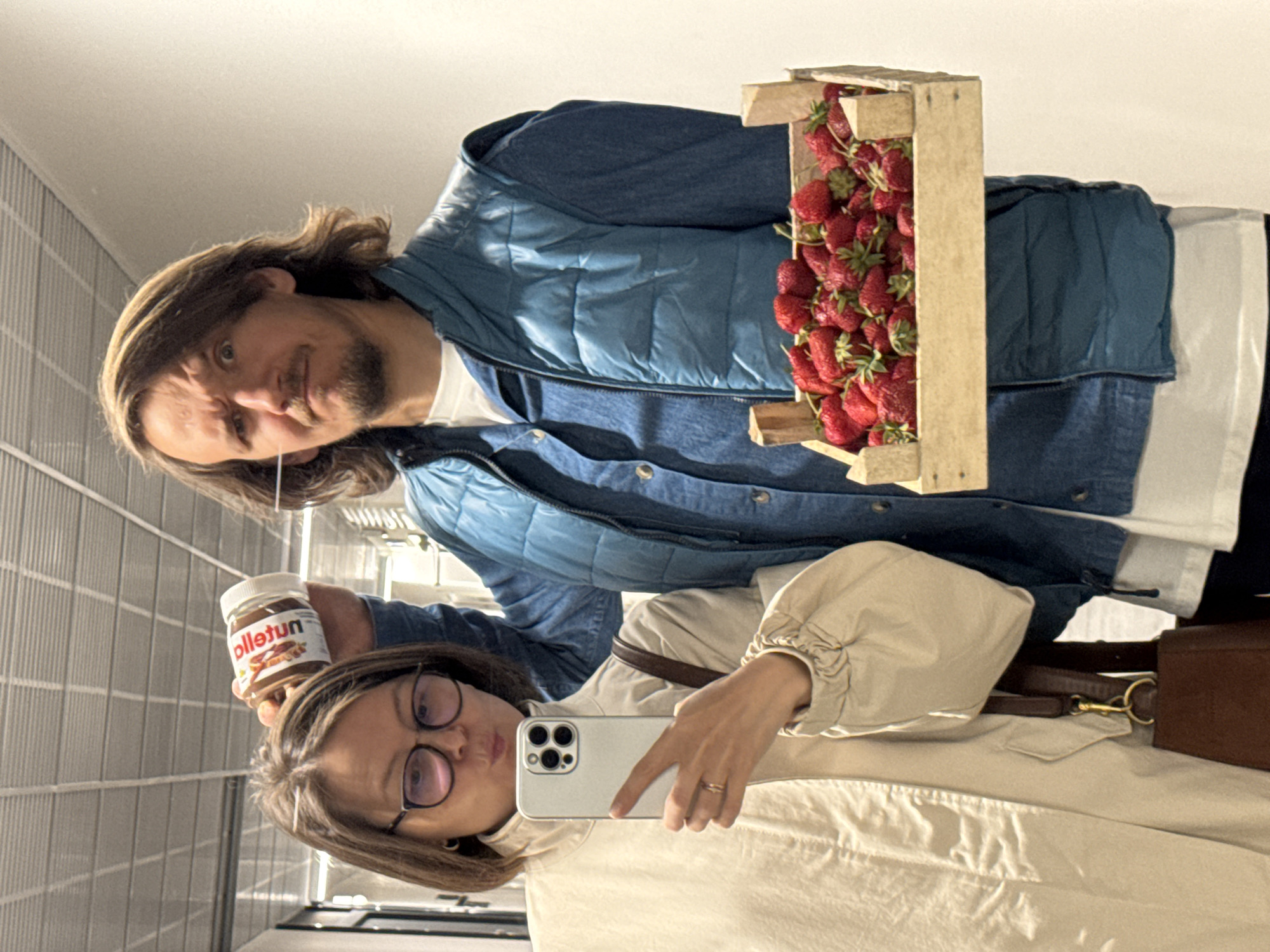 The image shows two people taking a selfie indoors. A woman with glasses holds a smartphone, pointing the camera towards herself and a man standing behind her to the left. In her left hand, she holds a jar labeled "ollełun", presumably a brand of nut butter. The man, who has long hair and a beard, holds a wooden crate filled with strawberries. He is wearing a blue puffy vest over a denim jacket. The woman is wearing a beige jacket with a brown purse slung across her body. The background appears to be a hallway with a metallic, vertically-lined wall. The city and country are not discernible from the image alone.