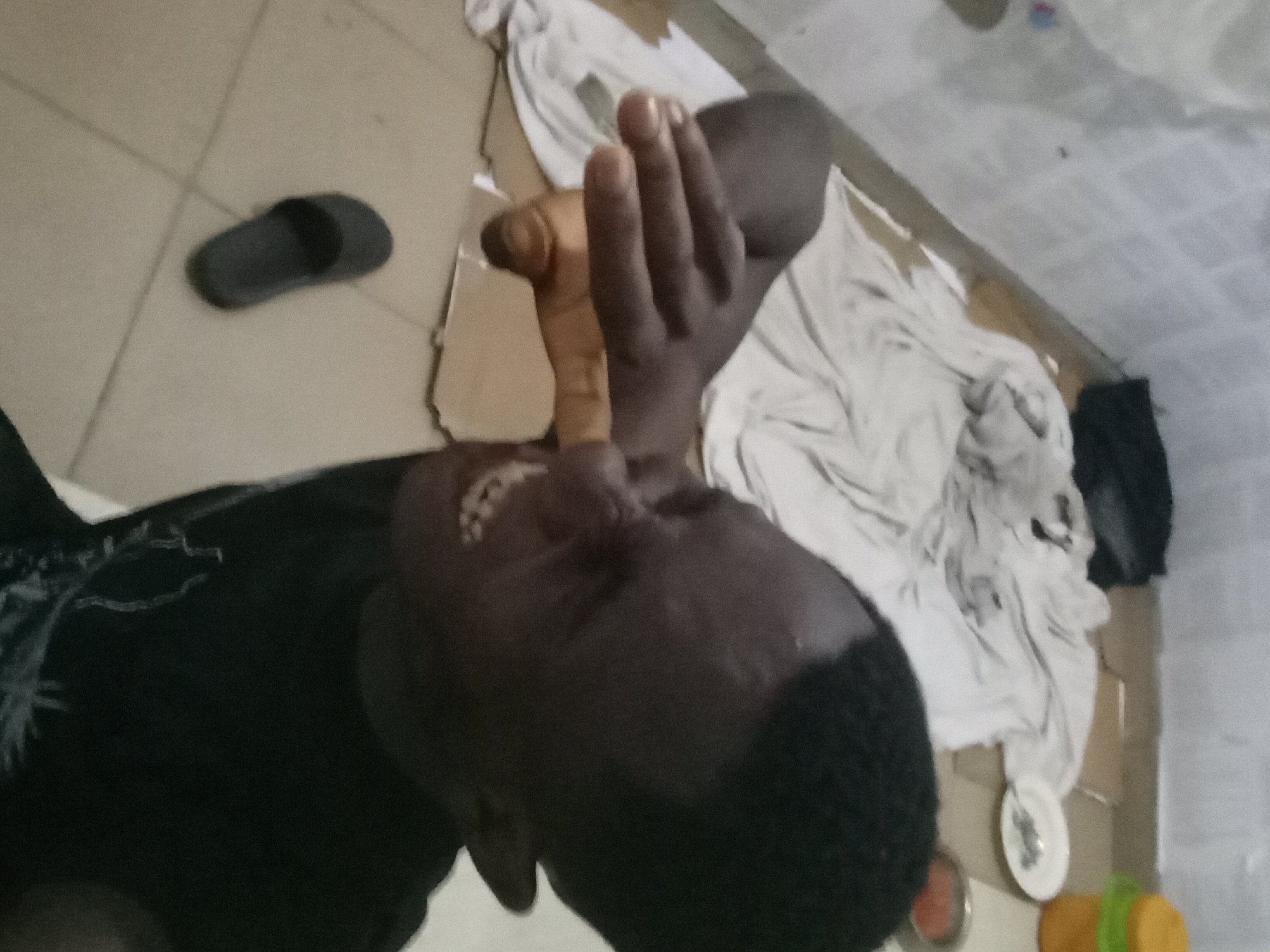 An overhead shot of a person with dark skin lying down on a surface covered with white fabric and cardboard. The person is wearing a black shirt. Their head is angled towards the camera, and they are holding a wooden cane in their right hand. A black sandal is visible on the tiled floor to the left. Near the fabric, a plate and a yellow container are visible. The location is Akwete, Nigeria.