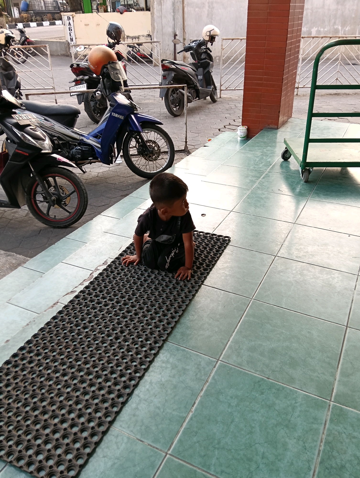 In Jaten, Indonesia, a young boy is sitting on a black, perforated mat placed on light green tiled flooring. He is wearing a dark t-shirt and dark pants. To his left, a black and blue Yamaha motorcycle is parked. Behind the motorcycle, several other motorcycles and a parked orange helmet are visible, suggesting a parking area or a street. Further in the background, a metal fence and buildings are present. To the boy's right, a green metal trolley with wheels is positioned on the tiled floor. The lighting suggests it is daytime, with sunlight casting shadows.