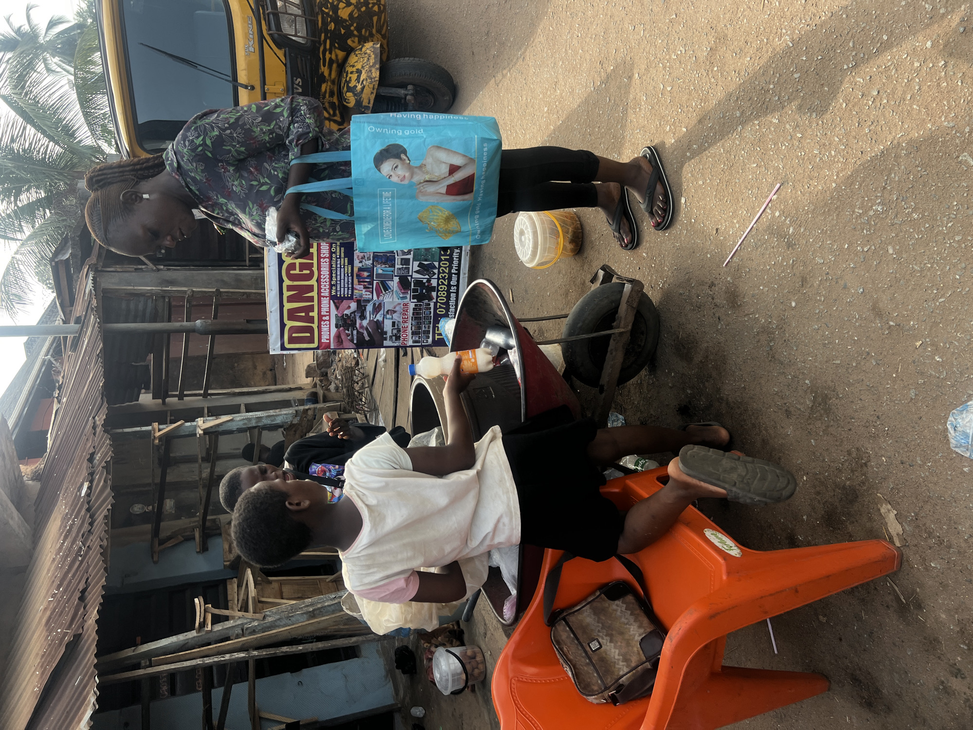 This medium shot captures a lively street scene in Obosi, Nigeria, under bright daylight. In the foreground, a woman stands, looking down at something in her hands. She wears a floral-patterned top and black leggings, and carries a prominent blue reusable shopping bag adorned with an image of a woman wearing jewelry and text such as "Owning gold Having happiness" and "LOVE SOMETHING FOR A LIFETIME." Her feet, in black flip-flops, show pink polished toenails.
To her right, another woman is seated casually on an orange plastic chair, holding a white bottle containing an orange liquid. A brown and black sling bag rests on the chair beside her, which also bears the brand name "Portune". In the background, a yellow commercial tricycle, commonly known as a keke napep, is partially visible, displaying "TVS" and "KING" branding.
Between the two women, a vertical banner advertises "DANG PHONES & PHONE ACCESSORIES SHOP," listing services like "PHONE REPAIR" and stating, "We Specialize On" with further, partially legible text, and "Satisfaction Is Our Priority." A contact number, "TEL 07089232013," is clearly visible. The surrounding environment features buildings with corrugated metal roofing and walls, typical of a bustling local commercial area. Scattered on the ground are white plastic containers, a red metal cart, and a light pink straw. Palm trees are visible in the upper left, adding to the tropical setting. The scene portrays everyday life and commerce in the area.
