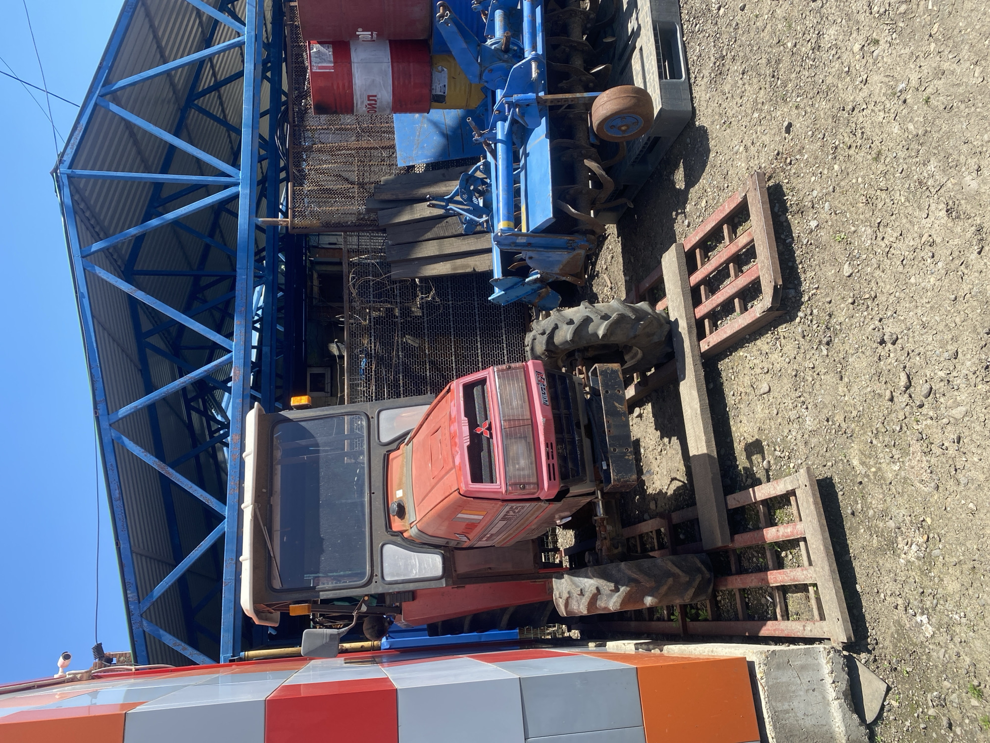 The image depicts a red tractor with a blue implement parked under a blue metal-framed structure in Neftyanik, Russia. The ground is dirt and gravel. A red and white building structure is visible in the foreground.