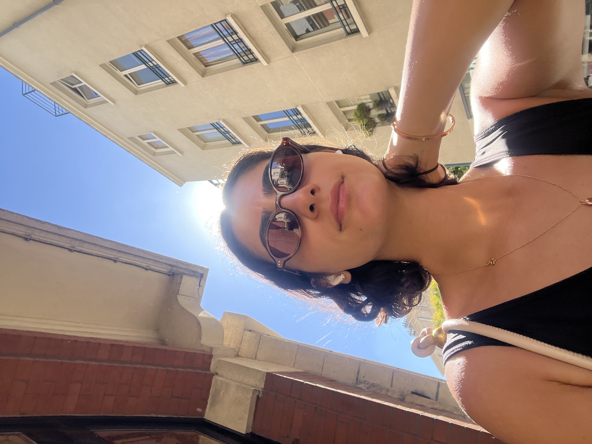 In a low-angle, sun-drenched selfie taken outdoors in Paris, France, a young woman with dark hair is captured. She is wearing sunglasses and appears to be looking upwards, with a faint smile on her lips. A delicate gold bracelet adorns her wrist, and a thin gold necklace with a small charm rests on her collarbone. She is wearing a black bikini top with a white trim. The background features the facade of a cream-colored building with multiple windows, some of which have dark metal bars. The sky is a bright, clear blue, and the sun is shining intensely, creating lens flare and highlighting the woman's skin with a warm glow. The bottom of the frame shows a brick wall. The overall impression is one of a casual, sunny day.