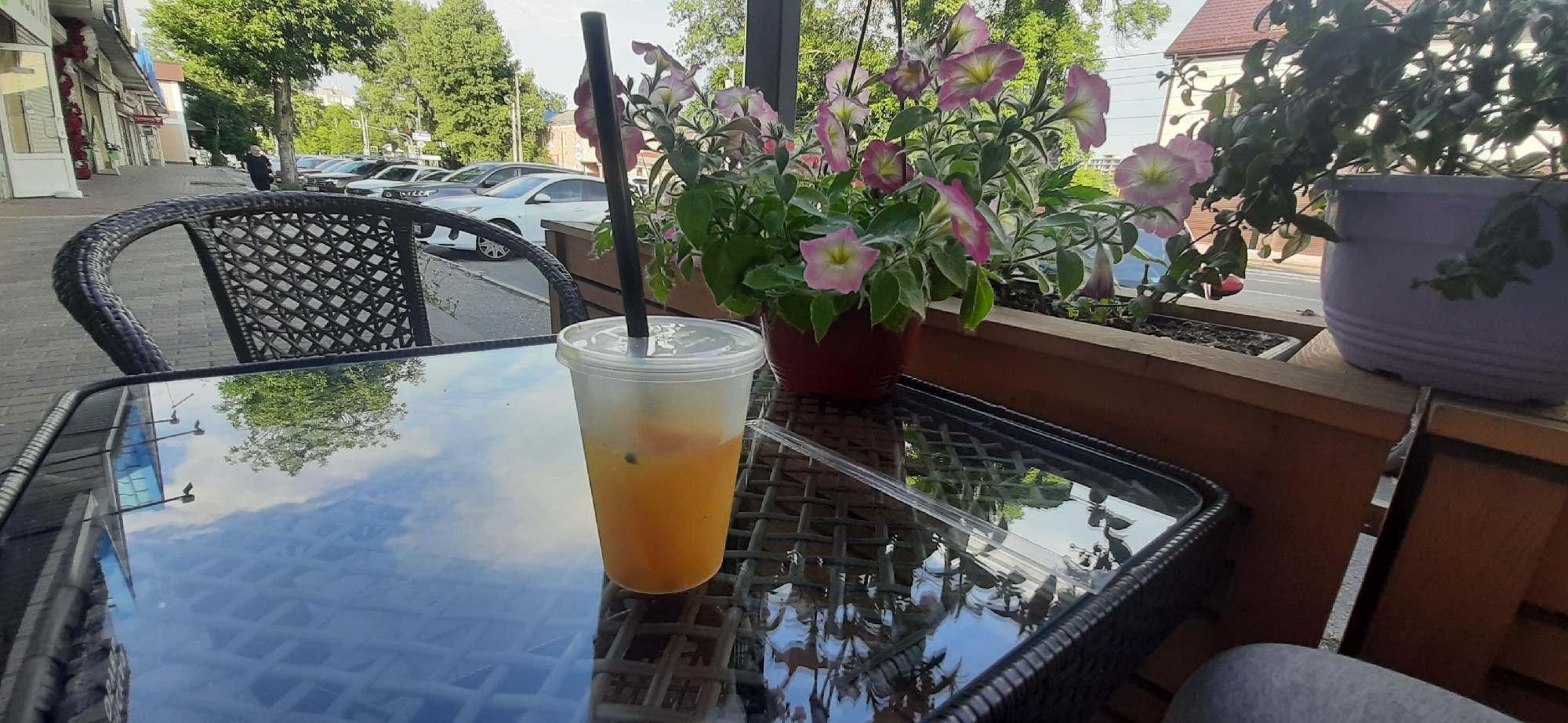 A clear plastic cup filled with an orange beverage and a black straw sits on a glass-topped table. The cup is positioned on the right side of the table, and it appears to be a refreshing drink. The table has a reflective surface, showing a mirrored image of the sky and trees. In the background, a black wicker chair is visible, partially obscuring the view of a street lined with parked cars and buildings. A woman wearing a dark headscarf and clothing is walking away from the viewer down the sidewalk. The scene is set outdoors, likely in a cafe or restaurant patio, given the table and chair. The presence of potted flowering plants, one with pink and white petunias, adds a touch of nature and vibrancy to the setting. The lighting suggests it is daytime, possibly late morning or afternoon, with soft sunlight filtering through the trees, creating a pleasant and relaxed atmosphere. The location is Yessentuki, Russia. There is no visible text in the image.