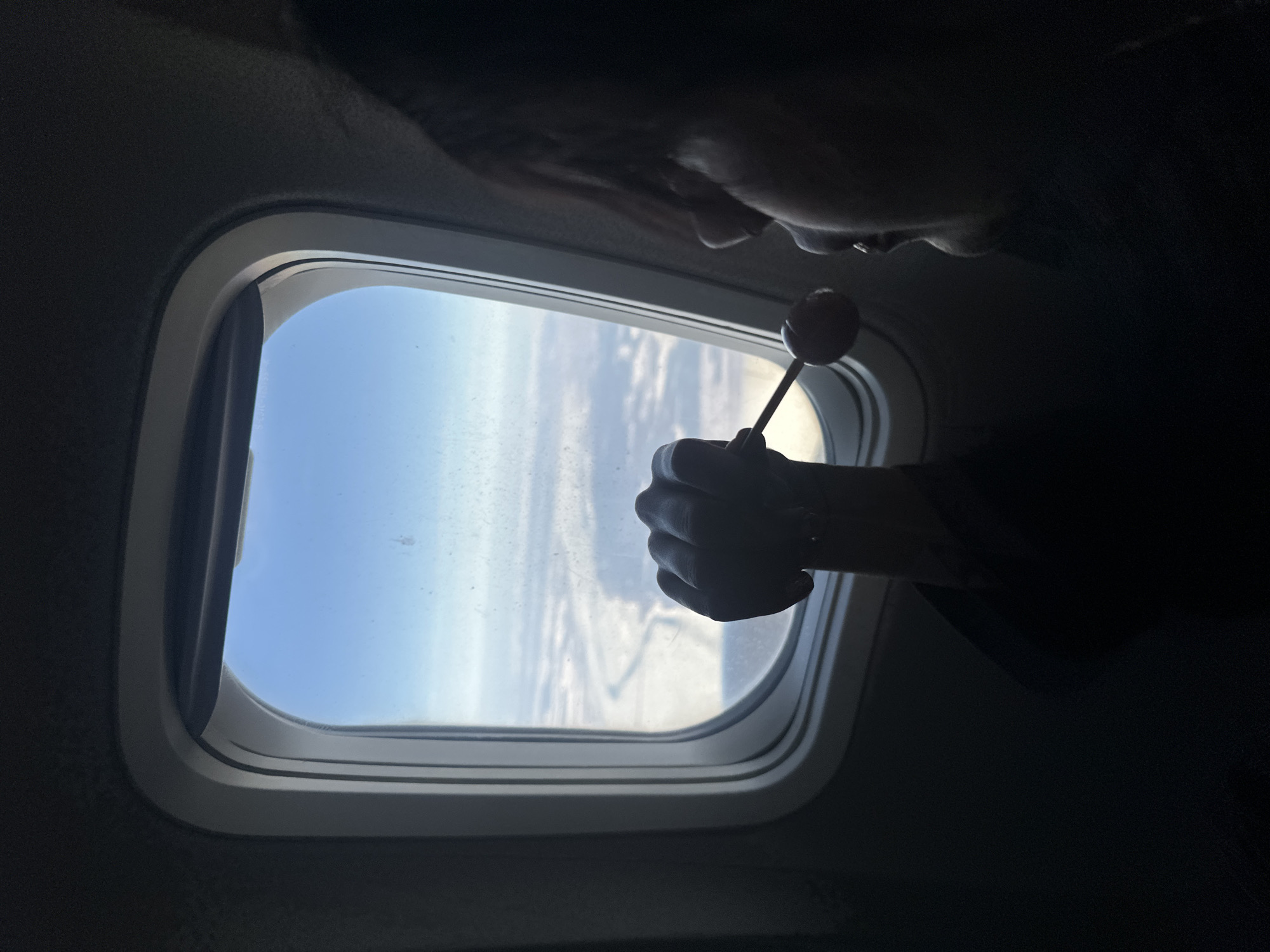 The image shows a person inside an airplane, looking out the window. The person is holding a lollipop in their hand, with the lollipop positioned in front of the window. Outside the window, the view consists of the sky and clouds, suggesting the plane is in flight. The interior of the plane is relatively dark. There is no information about the location in the city of Albaba, Russia.