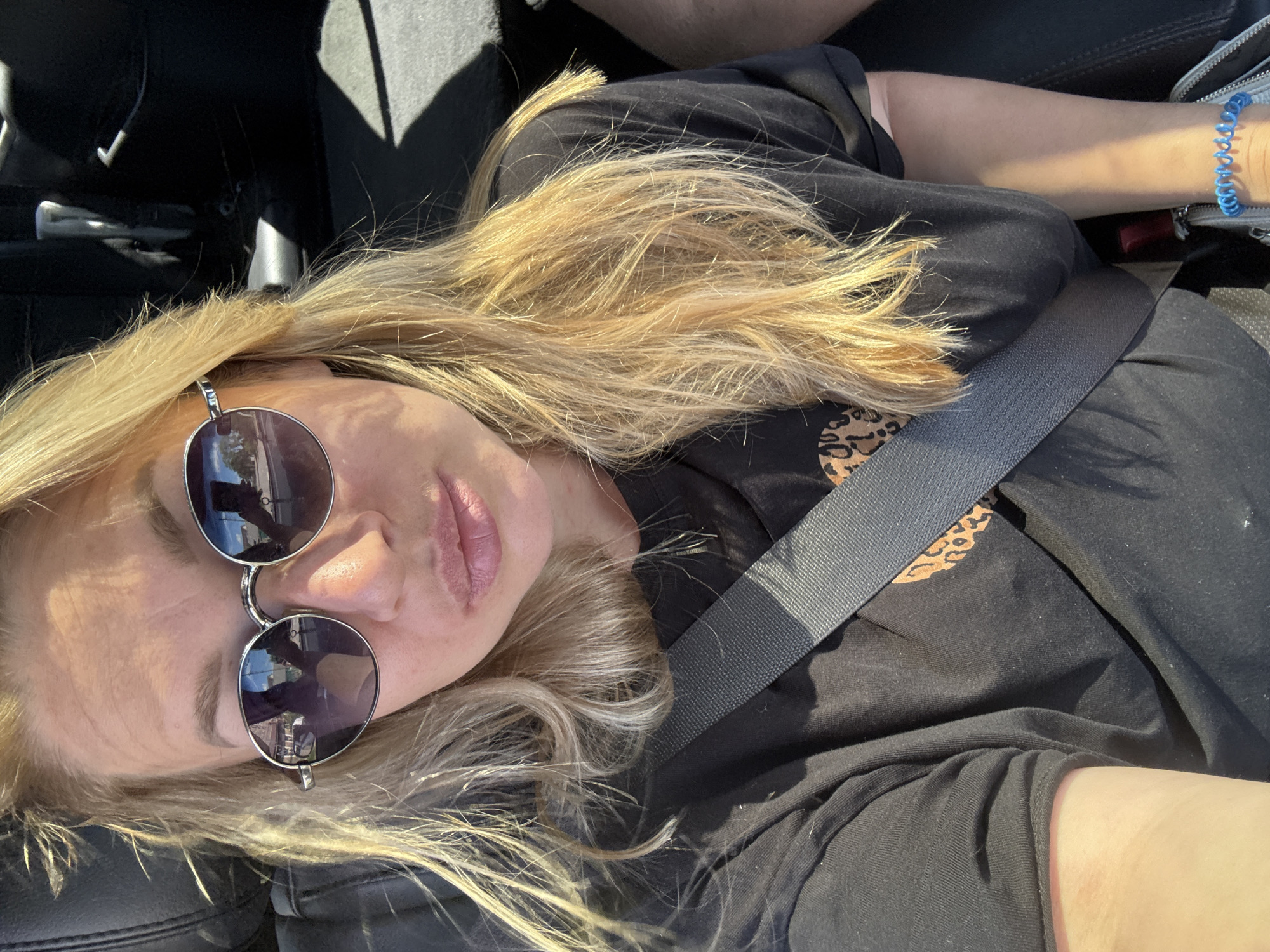 A woman with blonde hair, wearing sunglasses and a black t-shirt, is seen from the chest up, seated in a car. She has on a grey seatbelt and a blue bracelet. The background shows a dark car interior and the sunlight streaming in. The image is a selfie taken inside a vehicle. No specific location in Valday, Russia is identifiable.