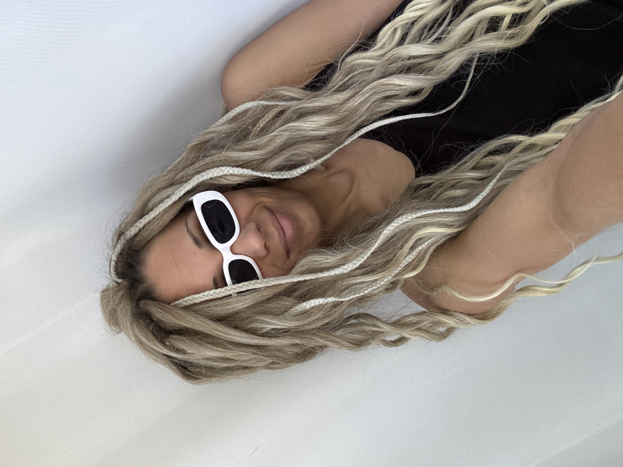 A woman with blonde braids is lying down, wearing white-framed sunglasses with black lenses and a black top. She is smiling slightly. The background is a plain white surface. The location cannot be determined to be in Rusanovo, Russia, or any specific city based on the image alone.