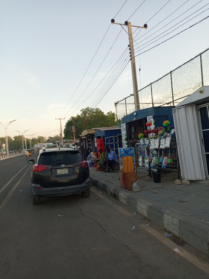 The image depicts a street scene in Maiduguri, Nigeria, located at coordinates 11.827 latitude and 13.155 longitude. A black SUV is parked on the side of the street, facing away from the camera. The street is paved and has a single lane of traffic moving in each direction. There are two yellow lines in the middle of the road that separate the lanes. The sidewalk to the right of the street is lined with a series of small, brightly colored shops selling various goods. There are several people sitting in front of the shops, seemingly engaging in conversations and/or transactions. A few trees are visible in the background, and a metal fence separates the road from the shops. Power lines run above the street, attached to a tall power pole. The sky is a light blue, and the overall scene seems to be in the late afternoon.