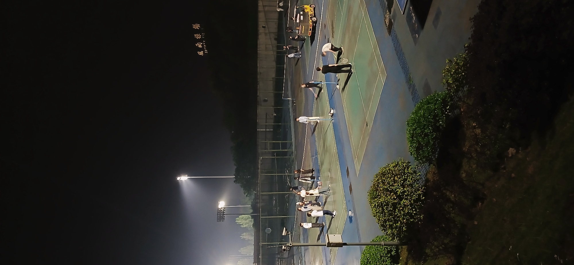 The image shows an outdoor badminton court located in Chongqing High-tech Zone, China at night. Several people are playing badminton on the court under artificial lighting. The court is blue and green with white lines, and there are green bushes along the side. Fences surround the court. In the background, there are trees and a building with writing in Chinese.