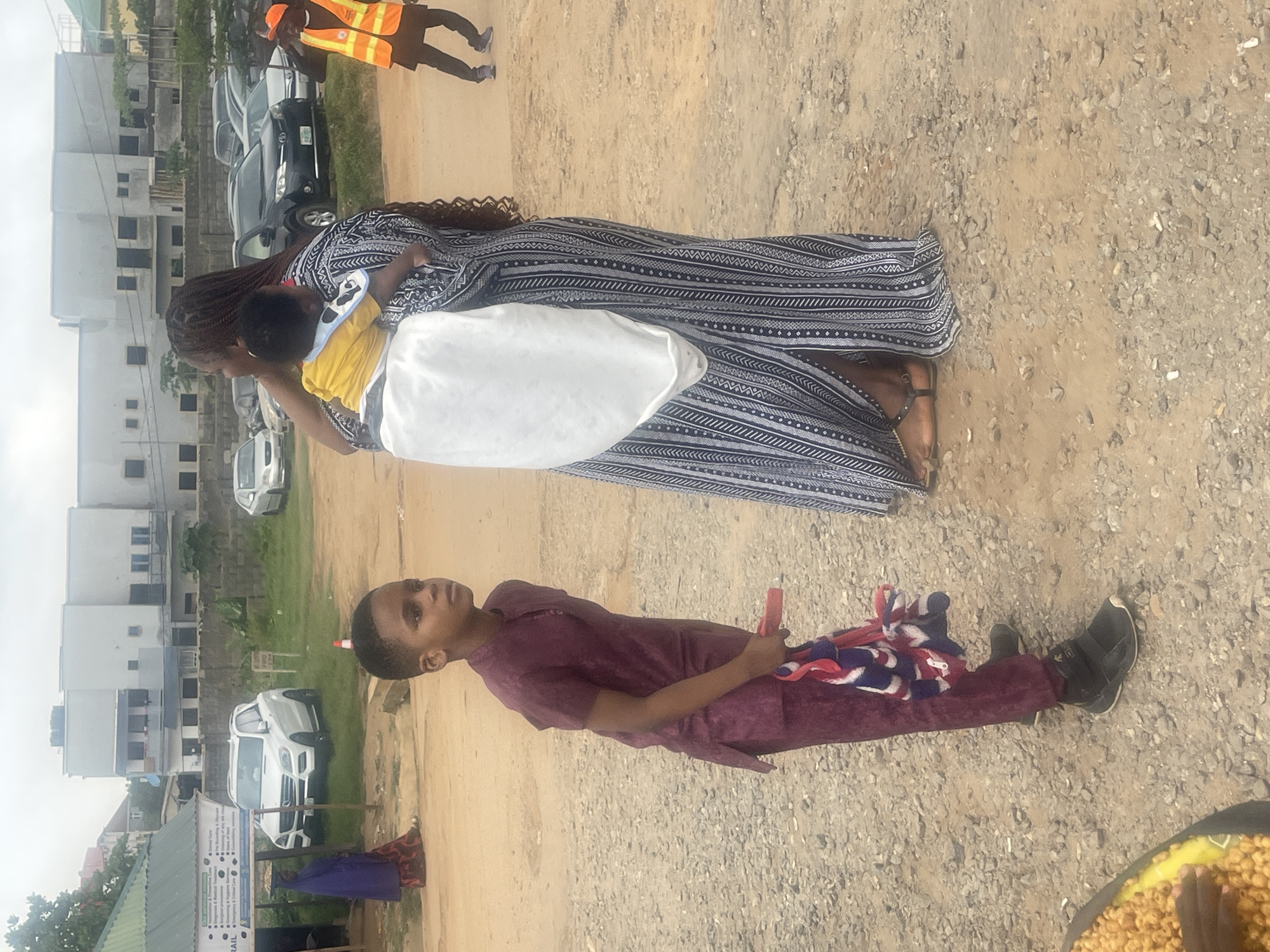 In Galadimawa Village, Nigeria, on a gravelly, dusty ground, a young boy in a maroon traditional outfit stands to the left of the frame, holding a bundle of red, white, and blue fabric in his right hand. He is looking to his left. Behind him, a woman with braided hair wears a long, flowing black and white patterned dress and carries a baby on her back, wrapped in a white cloth. The baby is wearing a yellow shirt. Further in the background, several vehicles are parked, including a white SUV and a dark SUV. A person wearing a bright orange safety vest is visible in the upper right quadrant of the image, walking away from the camera. A building with multiple windows and a flat roof is situated in the background, suggesting an urban or semi-urban setting within the village. A green structure with signage is present on the far left, and a few trees and bushes are scattered throughout the scene. The sky is overcast.