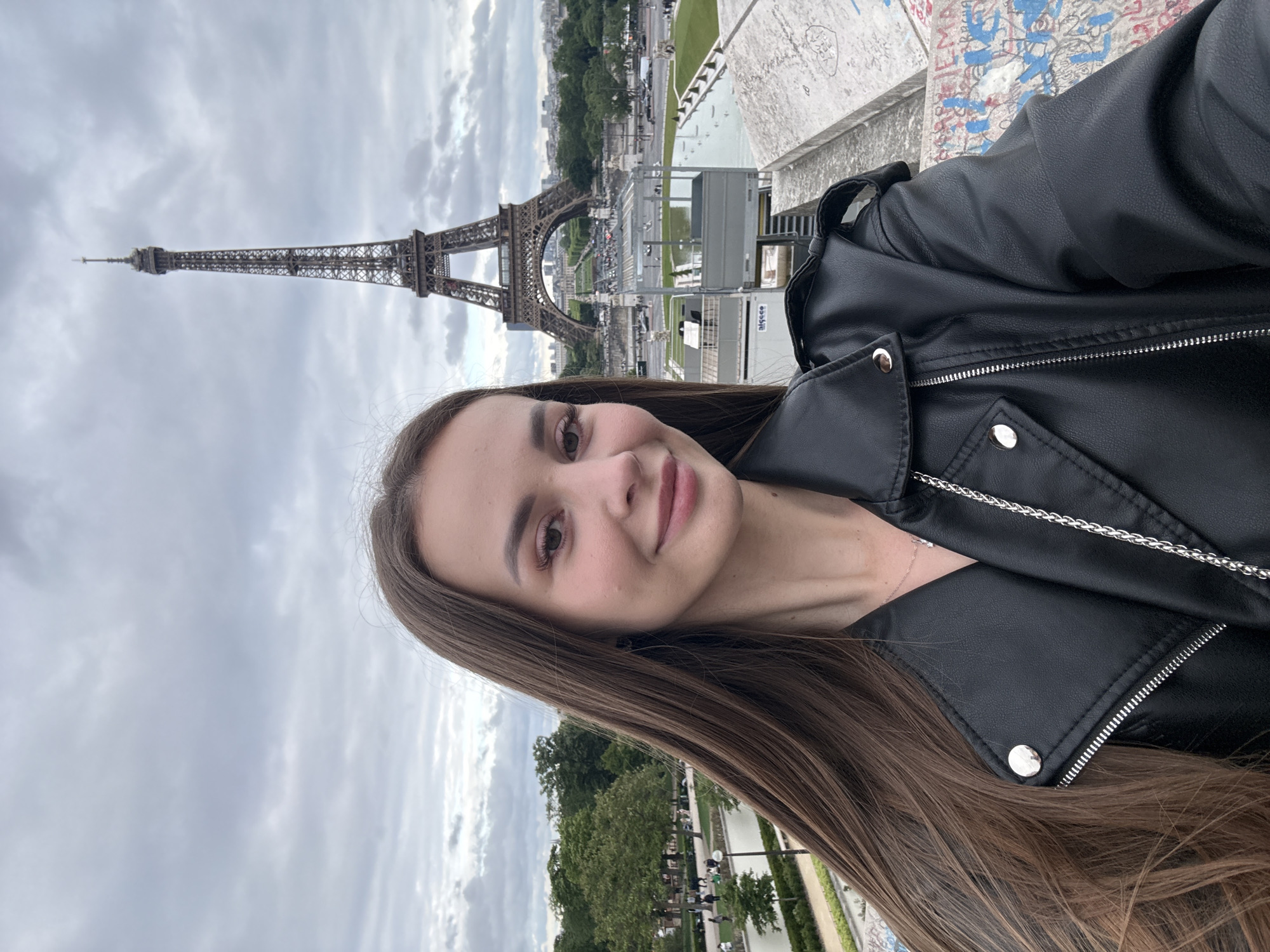 A vertical shot shows a woman in a black leather jacket standing in front of the Eiffel Tower in Paris, France. The tower looms in the background against a cloudy sky. Below the tower, trees and manicured grounds are visible. A graffiti-covered wall is to the upper-right of the image.