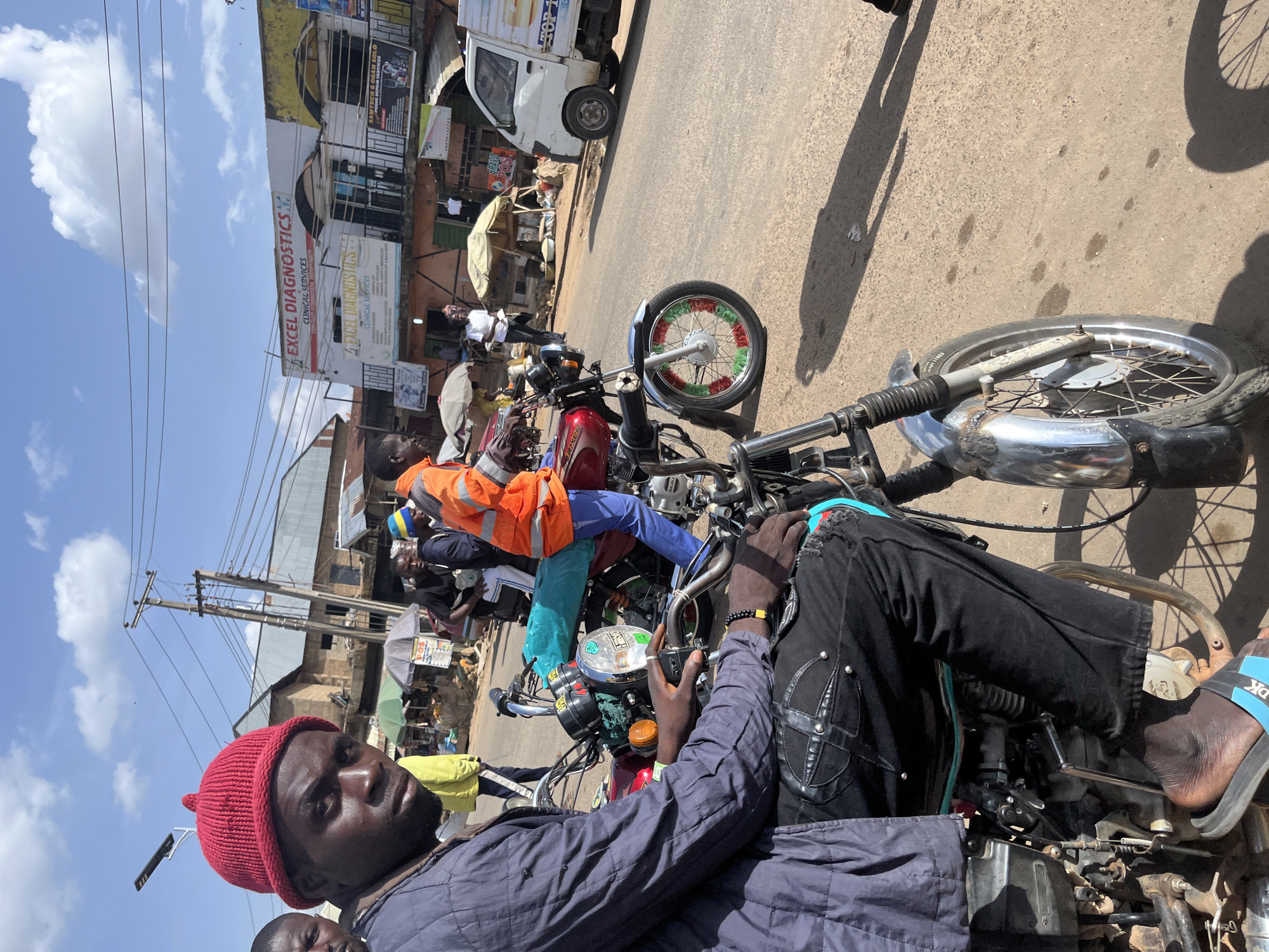 A medium shot captures a man wearing a red knitted beanie and a blue quilted jacket, sitting on a motorcycle in Gwong, Nigeria. He is looking directly at the camera with a serious expression. His black pants are decorated with a criss-cross pattern and silver studs. His right foot is visible, wearing a black and blue sandal. Another motorcycle, red and maroon, is parked next to him, with a turquoise bag on its seat. In the background, several other people are present, some wearing bright orange or yellow jackets, and a white truck is parked on the right side of the street. Buildings with signage, including "EXCEL DIAGNOSTICS," line the street, indicating a commercial area. The scene is set outdoors on a sunny day with a partly cloudy sky.