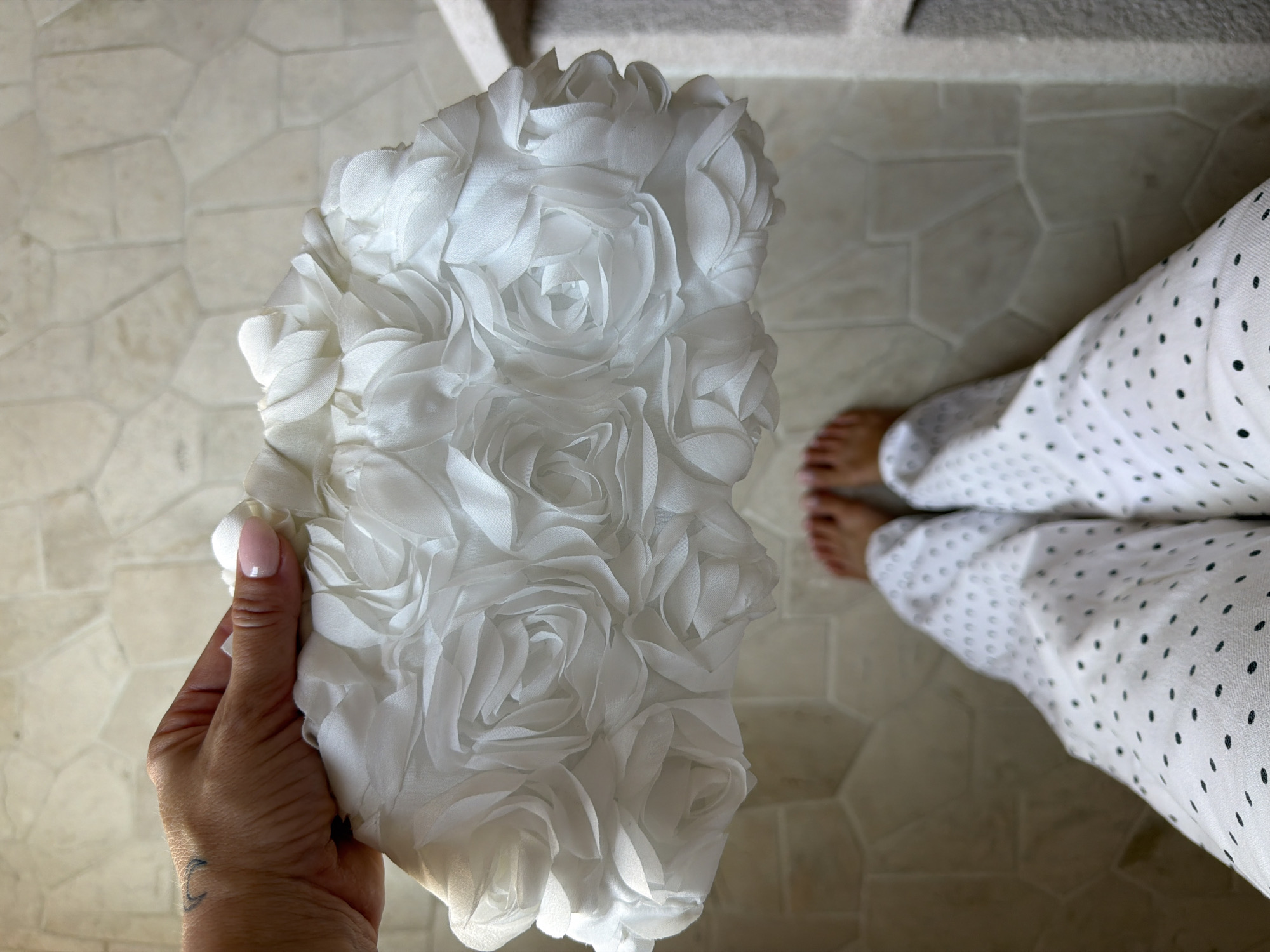 In Mengwi, Indonesia, a close-up, over-the-shoulder shot captures a woman's hand, adorned with a light pink manicure and a subtle tattoo on her wrist, holding a white clutch bag. The bag is embellished with numerous fabric rosettes, creating a textured floral pattern. Below, the woman's feet are visible, clad in white pants with a scattered black polka dot print, standing on a floor tiled with large, irregularly shaped beige stones. The scene is lit by natural light, casting soft shadows and highlighting the delicate details of the clutch.
