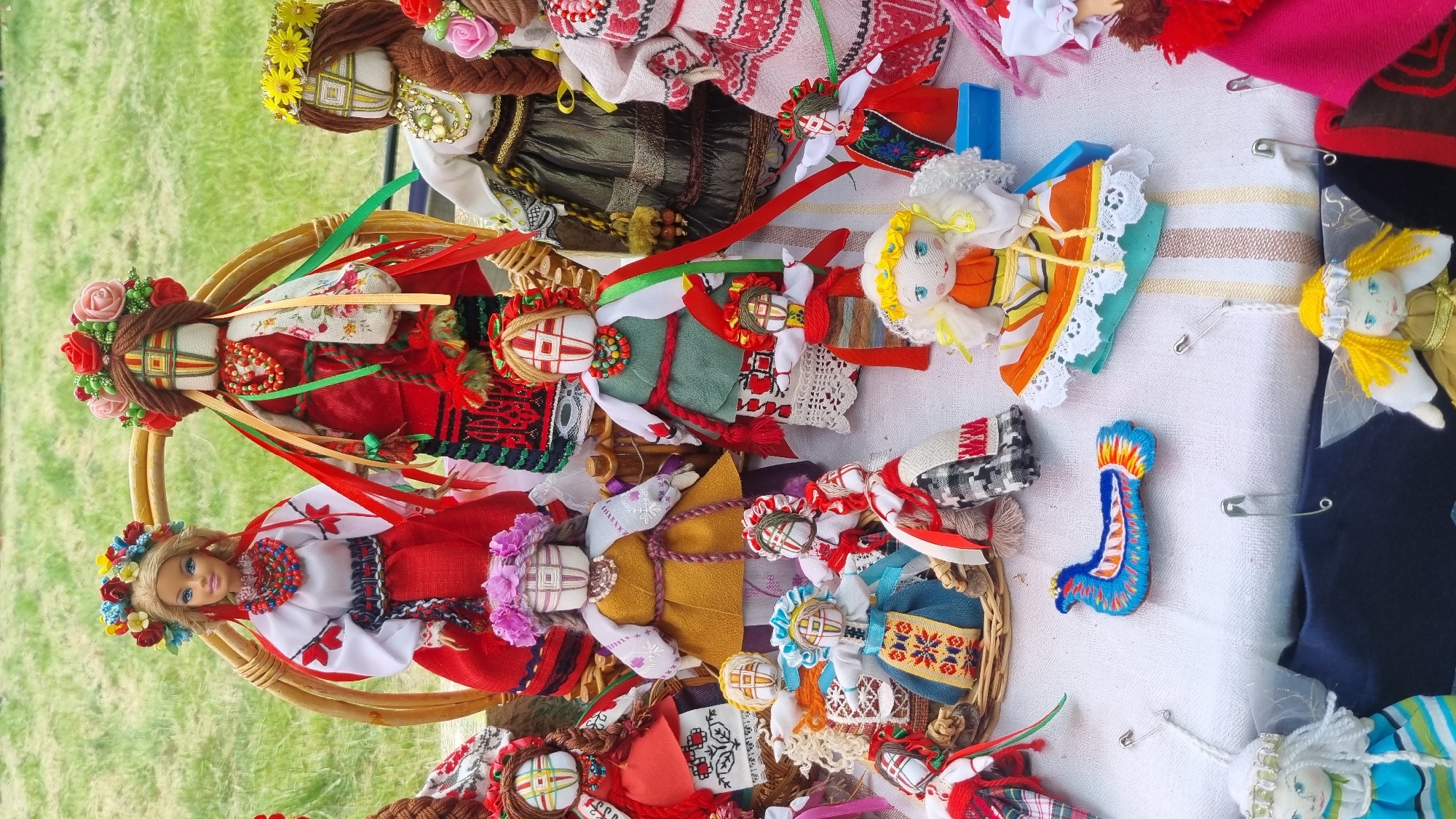 The image shows a collection of handmade dolls displayed on a white, striped cloth. The dolls vary in size and style, with many dressed in traditional Ukrainian folk costumes featuring embroidered patterns and colorful ribbons. Some dolls are adorned with floral wreaths and beaded necklaces. In the background, there's a glimpse of green grass, suggesting an outdoor setting. The dolls are arranged in a somewhat cluttered, artistic manner, some sitting in woven baskets, while others are placed directly on the cloth. In the center of the display, a small, blue embroidered bird figurine is prominently featured. The overall impression is that of a market stall or craft fair. While the image itself does not contain explicit references to Smila, Ukraine, the style of the dolls suggests a possible connection to Ukrainian folk art traditions.