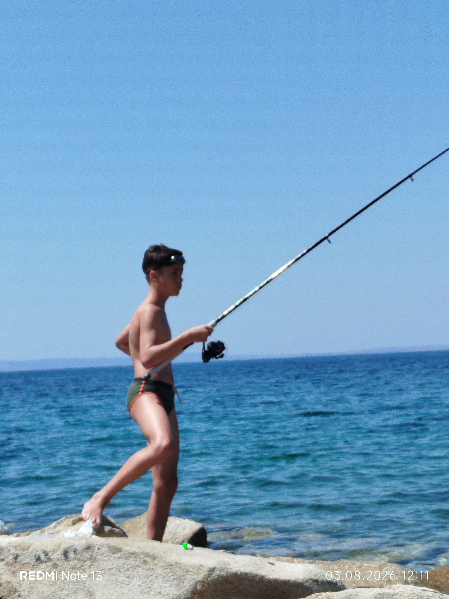 A young boy stands on a rocky shore, fishing with a rod. He is wearing swimming trunks and goggles on his forehead. The boy is looking out at the blue sea, which stretches to the horizon under a clear blue sky. The rocky shore is made up of large, light-colored boulders. The water is calm with small ripples. The overall scene is one of a peaceful day at the beach in Sithonia, Greece.