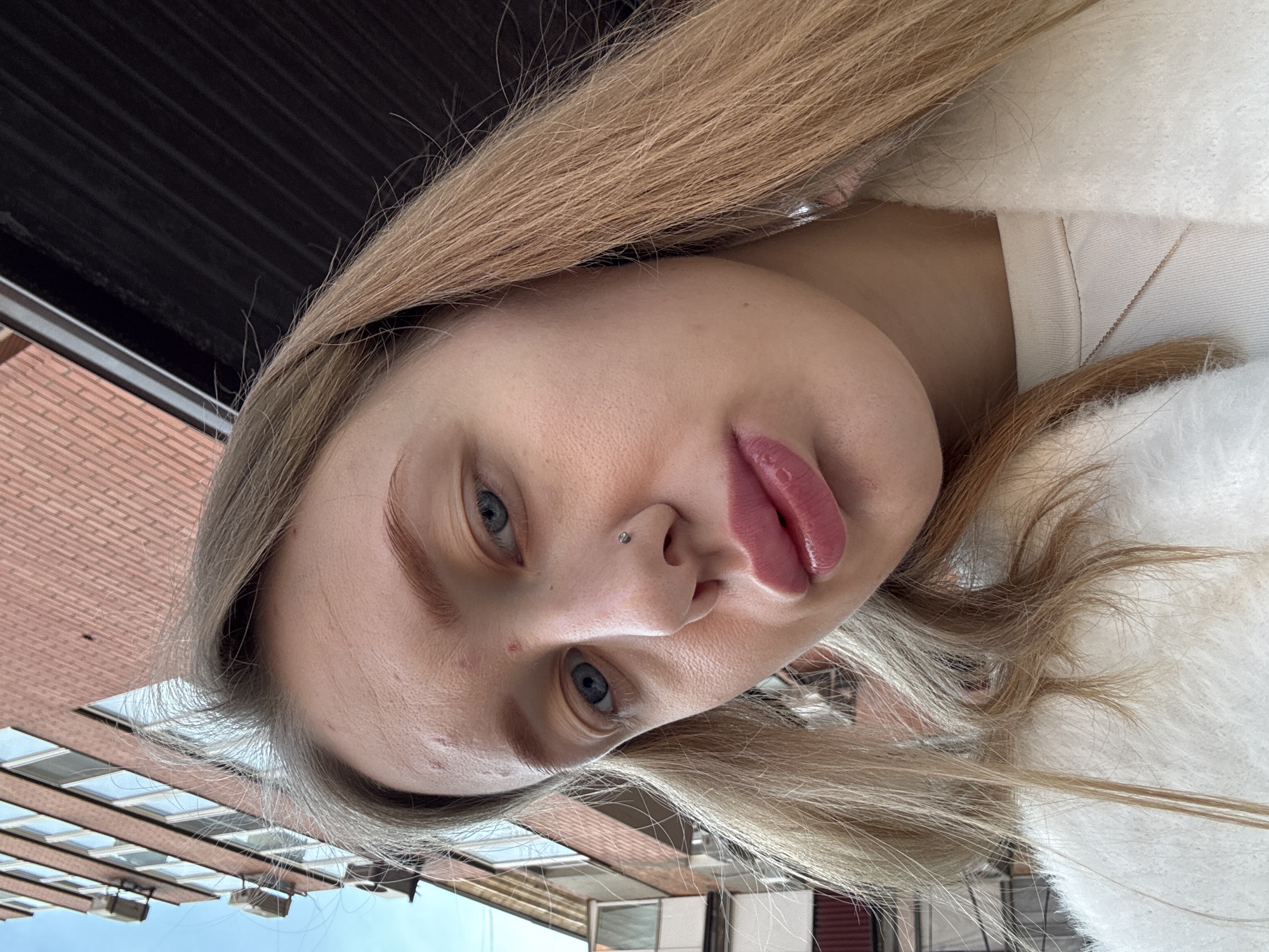 A young woman with blonde hair and blue eyes is looking directly at the camera. She has a nose piercing and glossy lips. The background shows a brick building with windows. The setting is likely outdoors in Rostov-on-Don, Russia, judging by the location metadata. The woman is wearing a white top.