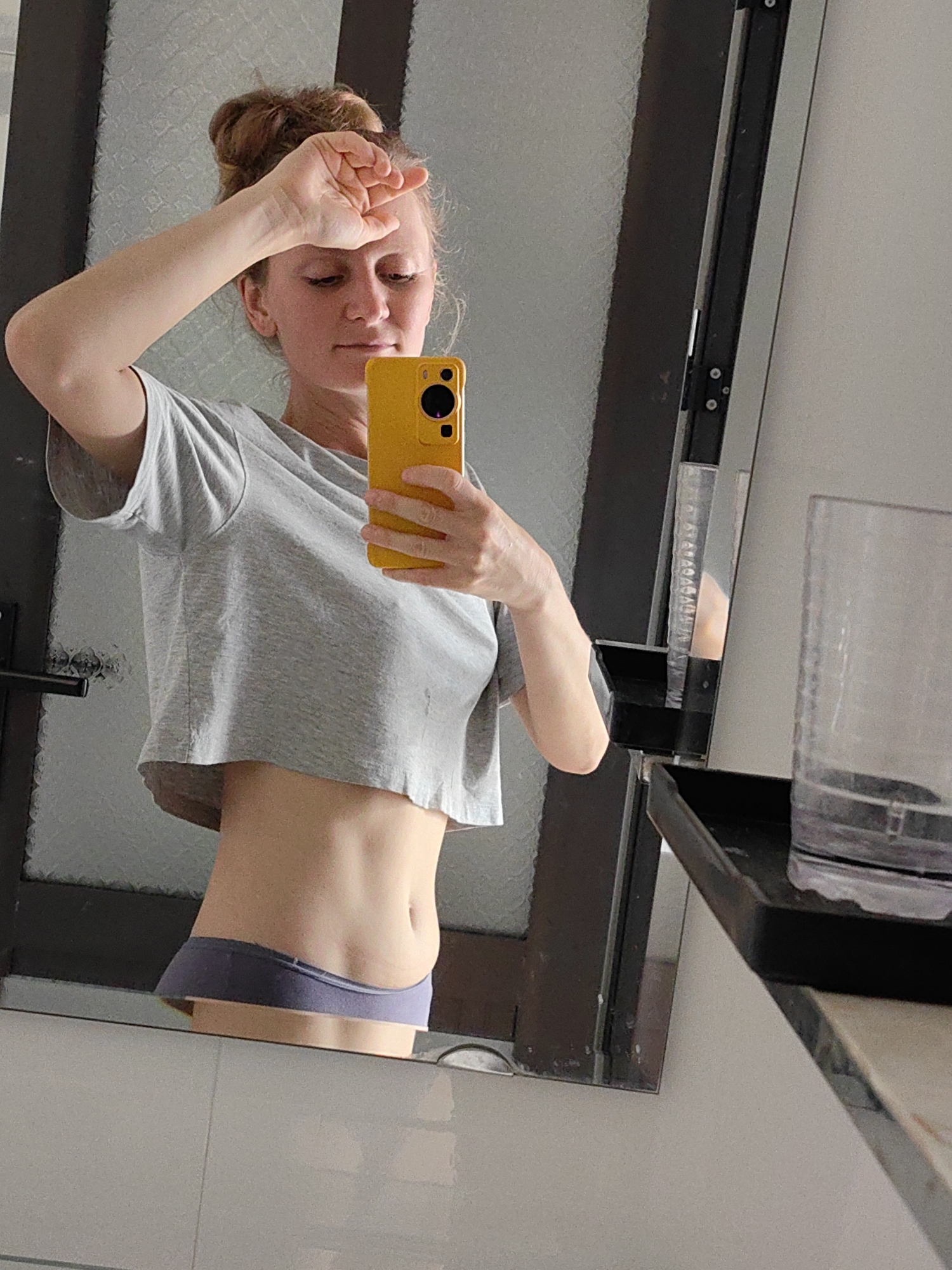 A woman is taking a selfie in a bathroom. She is wearing a gray crop top and blue underwear. She is holding a yellow smartphone in her right hand, partially covering her face, and her left hand is on her forehead. The bathroom features a mirror with a dark frame, a transparent glass near a black tray, and white tiles. The location appears to be indoors in a private residence or bathroom, not identifiable as Bai Chay Ward, Vietnam, based on visible information.