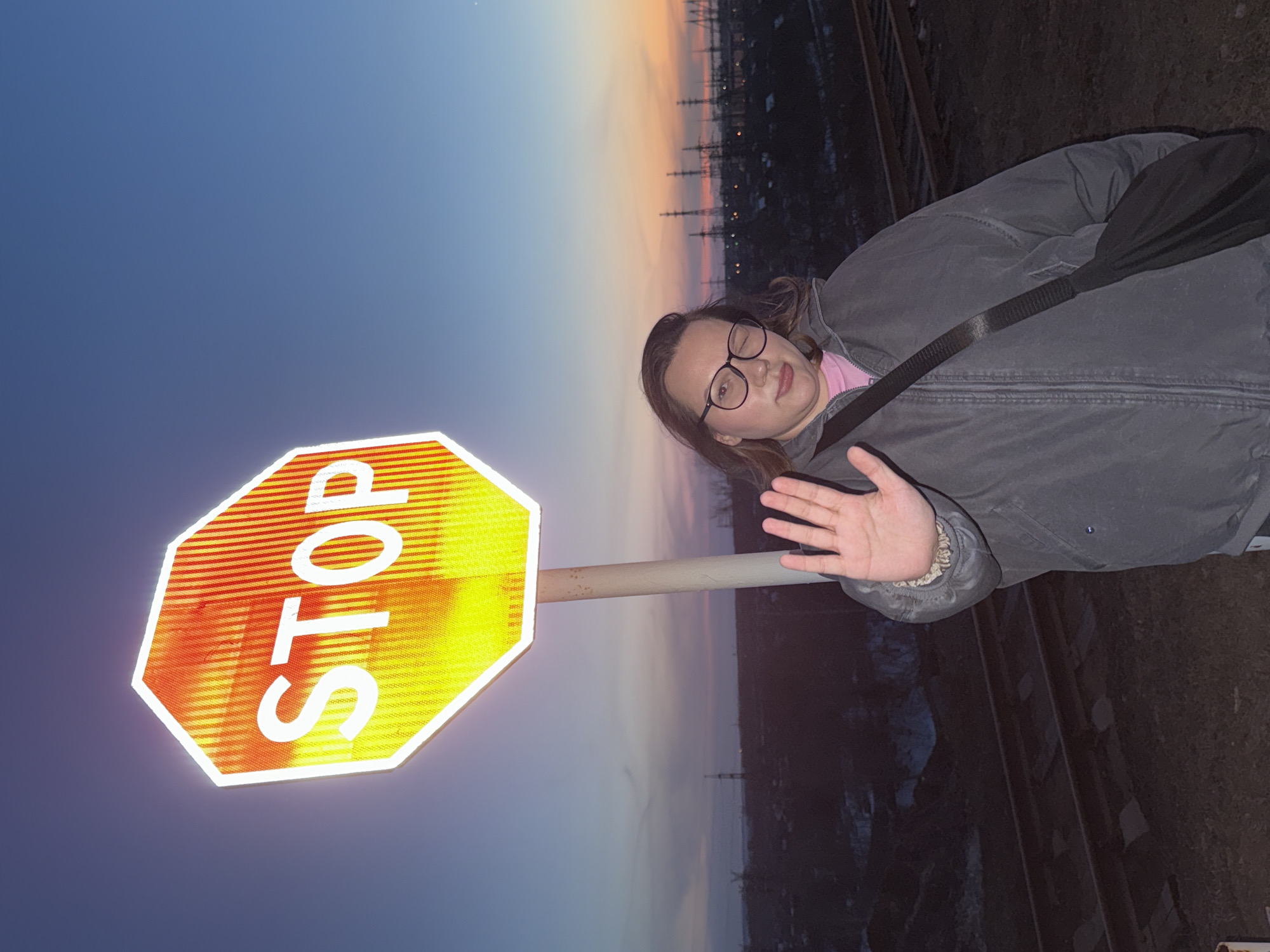 A vertical image shows a person and a stop sign against a sky at dusk. The person, wearing glasses and a gray jacket with a black strap across the chest, stands to the right of a yellow and red stop sign. The sign is on a pole, with the word "STOP" written vertically in white letters. The background shows the edge of what appears to be a railway track, with power lines visible in the distance under a multicolored sky. The picture was taken in the city of Chaikovsky, Russia.