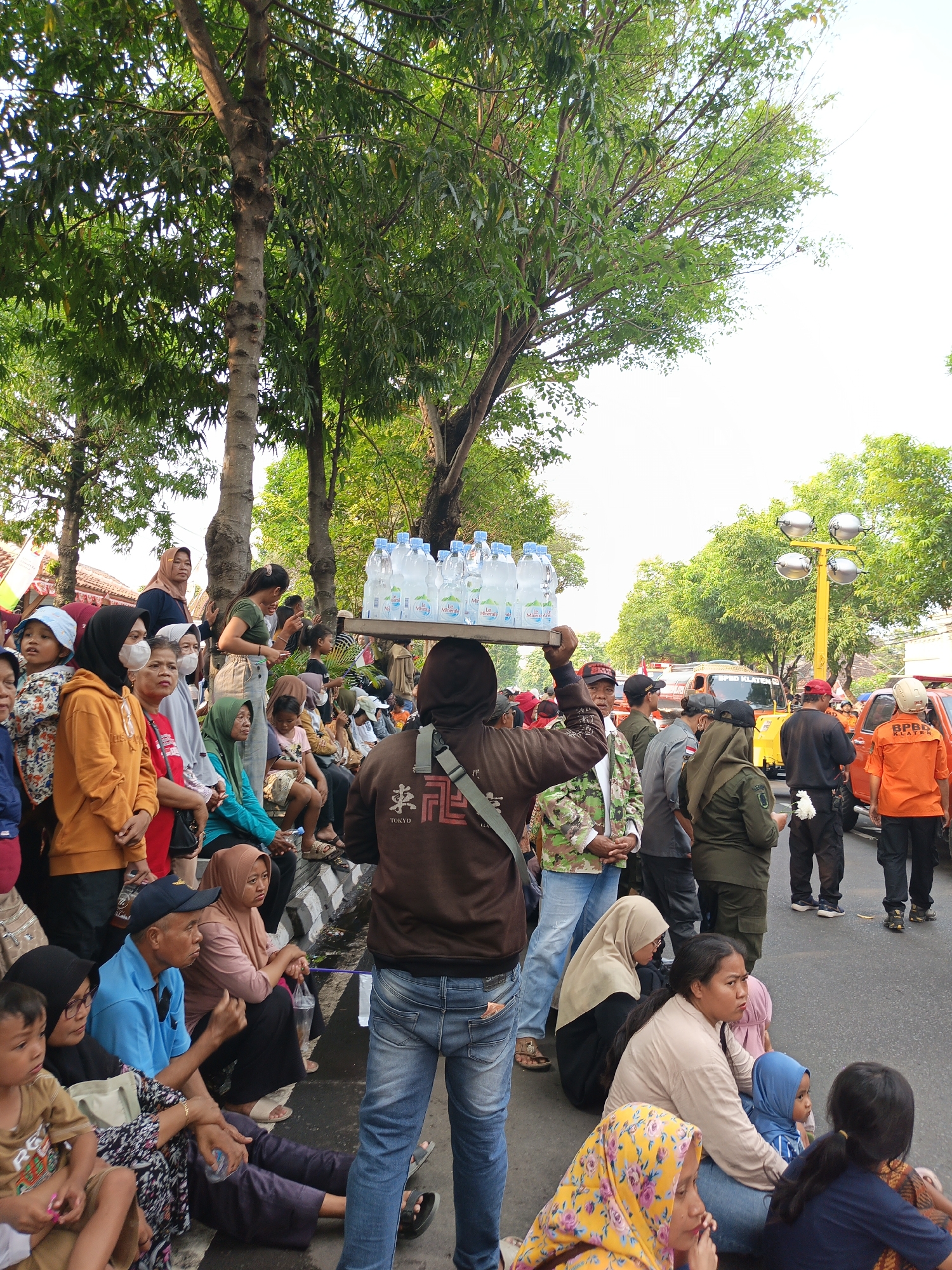 In Gayamprit, Indonesia, a crowd of people, including men, women, and children, are gathered outdoors, possibly for an event or festival. A man in the foreground carries a tray of bottled water on his head, navigating through the crowd. Many individuals are seated on the ground or on a low wall, while others stand. The environment features trees with lush green foliage, suggesting a warm climate. In the background, vehicles, including what appears to be a yellow emergency vehicle with "BPBD KLATEN" written on it, and an orange vehicle are visible, along with streetlights. The overall atmosphere seems to be one of communal gathering and perhaps an outdoor event.