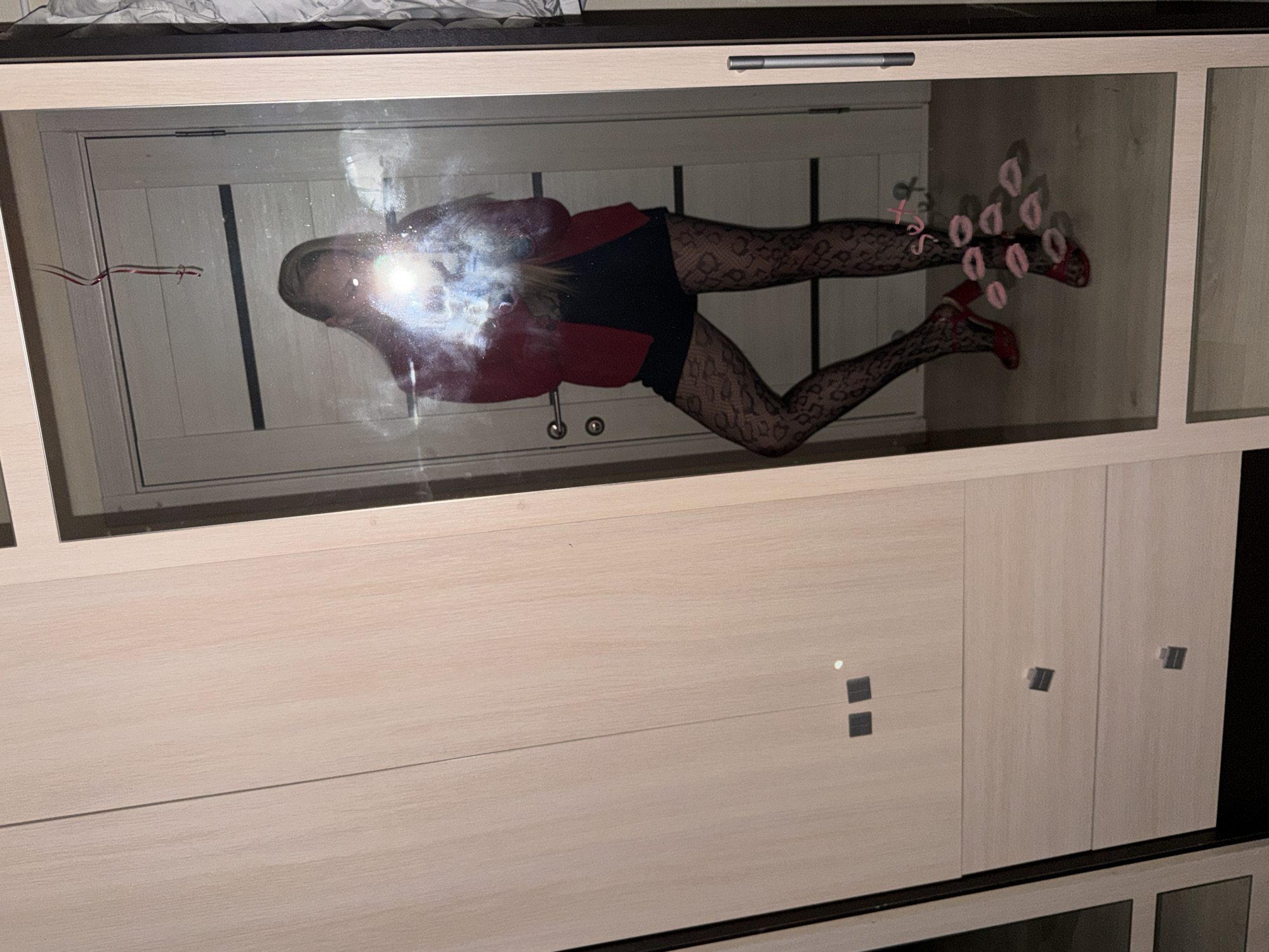 The rotated image captures the reflection of a person, likely female, in the glass doors of a wardrobe in Izhevsk, Russia. The subject is positioned horizontally across the frame, wearing a red top or jacket, black shorts or a short skirt, patterned fishnet stockings, and red high-heeled shoes. A bright flash or light source creates a strong glare over the upper part of their body, obscuring facial details.
On the glass surface, there are multiple pink lipstick marks shaped like kisses, and handwritten text that includes "XOXO" and "поцелуй" (Russian for "kiss"). A thin red and white string-like decoration is also affixed to the glass on the left. The background reflected in the glass appears to be an indoor setting with a light-colored door or wall.
Below the glass-fronted section, the wardrobe features light wooden panels with discreet square handles, indicating a modern, minimalist style. The overall scene suggests an intimate indoor setting, possibly a bedroom or dressing area. The lighting is artificial, implying the photo was taken in the evening or at night. The pose and the lipstick marks convey a playful and confident mood.