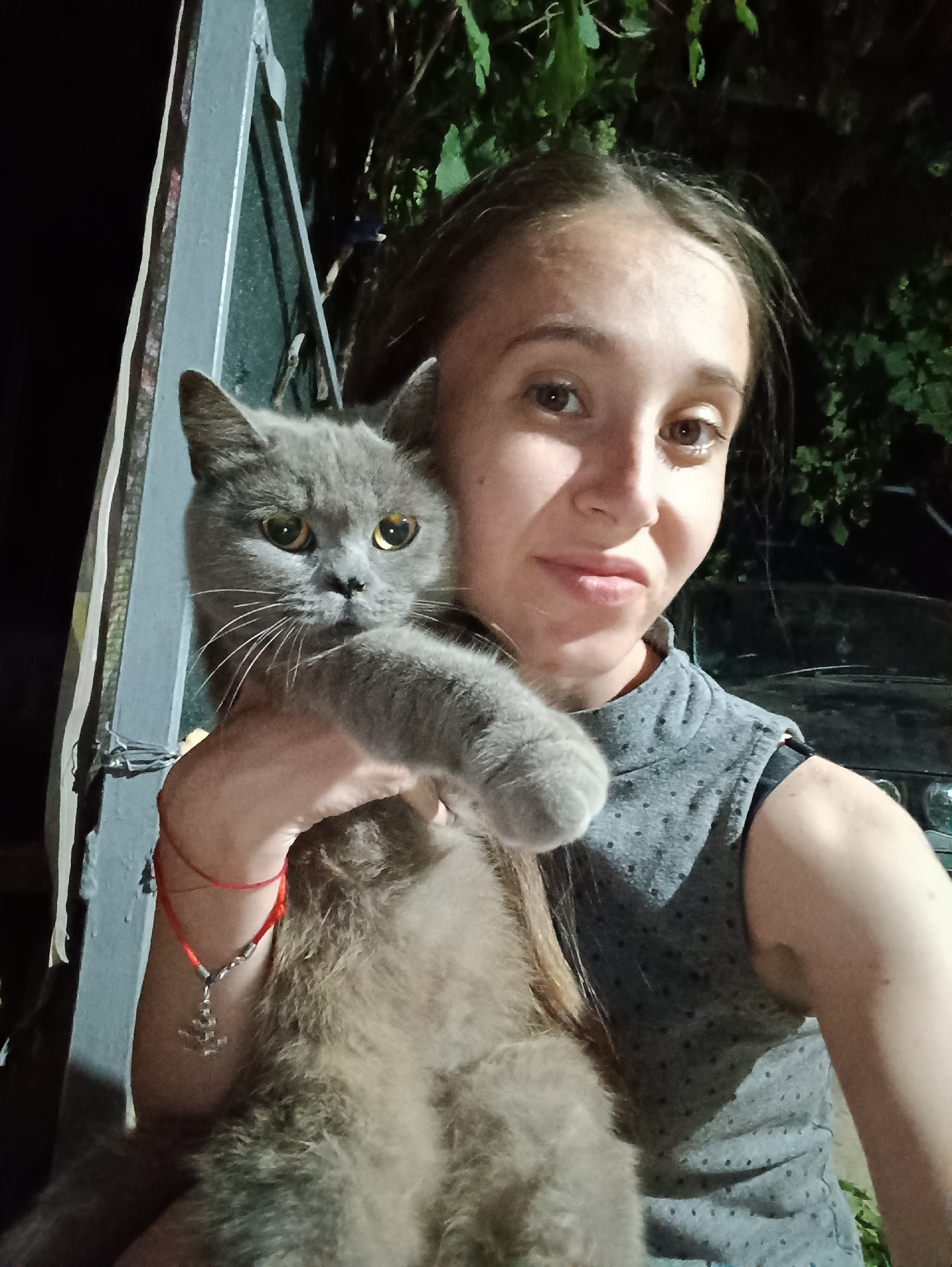 A young woman with long brown hair is holding a gray cat in front of her. She is wearing a gray sleeveless top with a high neckline and a black bra strap visible. The cat, with yellow eyes, is nestled in her arms. A red string bracelet with a silver anchor pendant is visible on her left wrist. The background is dark with some foliage, suggesting an outdoor setting at night. There is a metal structure to the left of the woman. The image appears to be a selfie. The location is Vypasne, Ukraine.