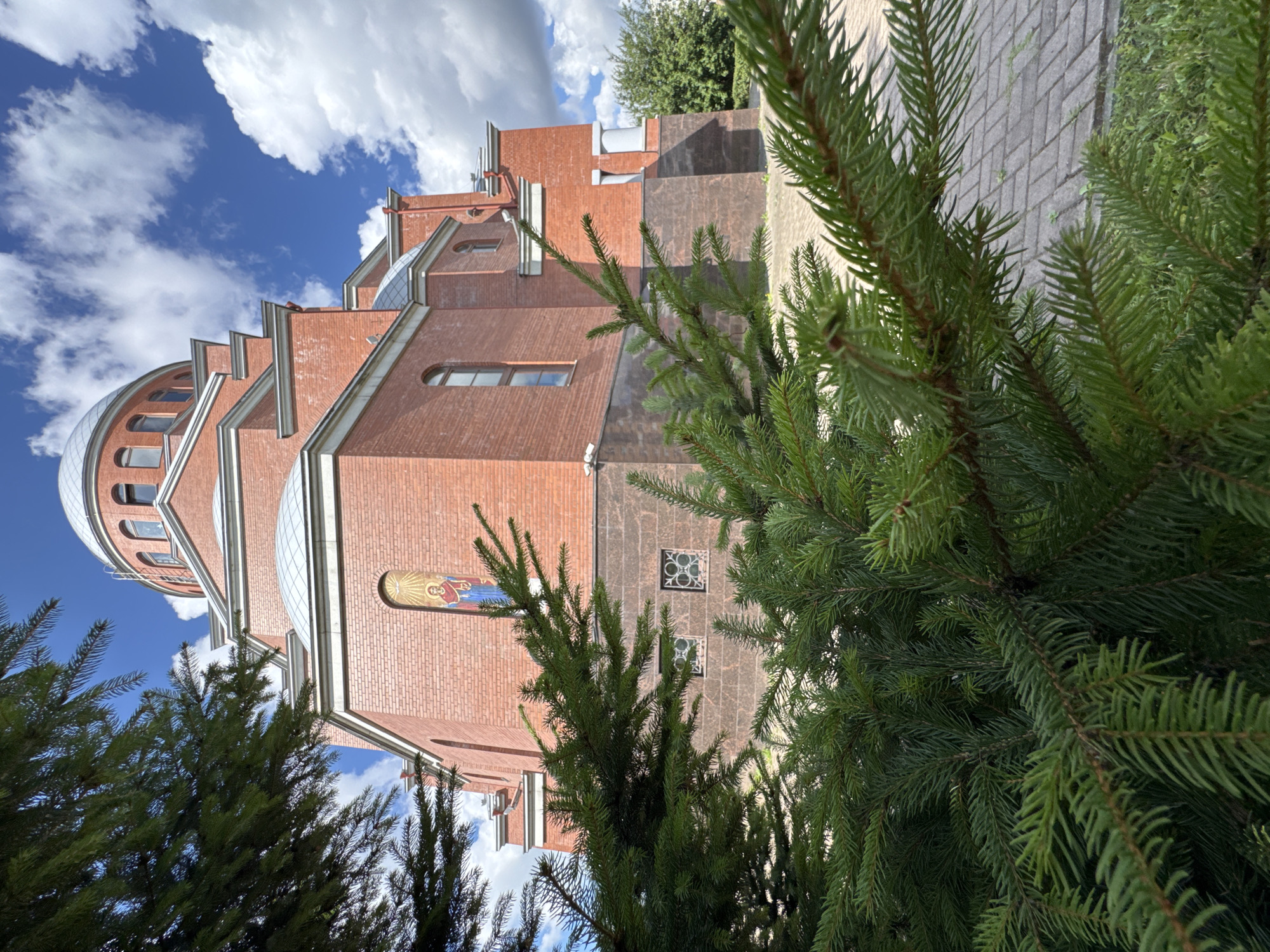 The image shows an exterior view of the Orthodox Church of the Resurrection of Christ in Sinyavino, Russia. The church is constructed of red brick with a domed roof. In the foreground, evergreen branches partially obscure the view of the church. The sky above is blue with scattered white clouds. A paved walkway is visible in the lower right portion of the image. There are no people visible in the image.
