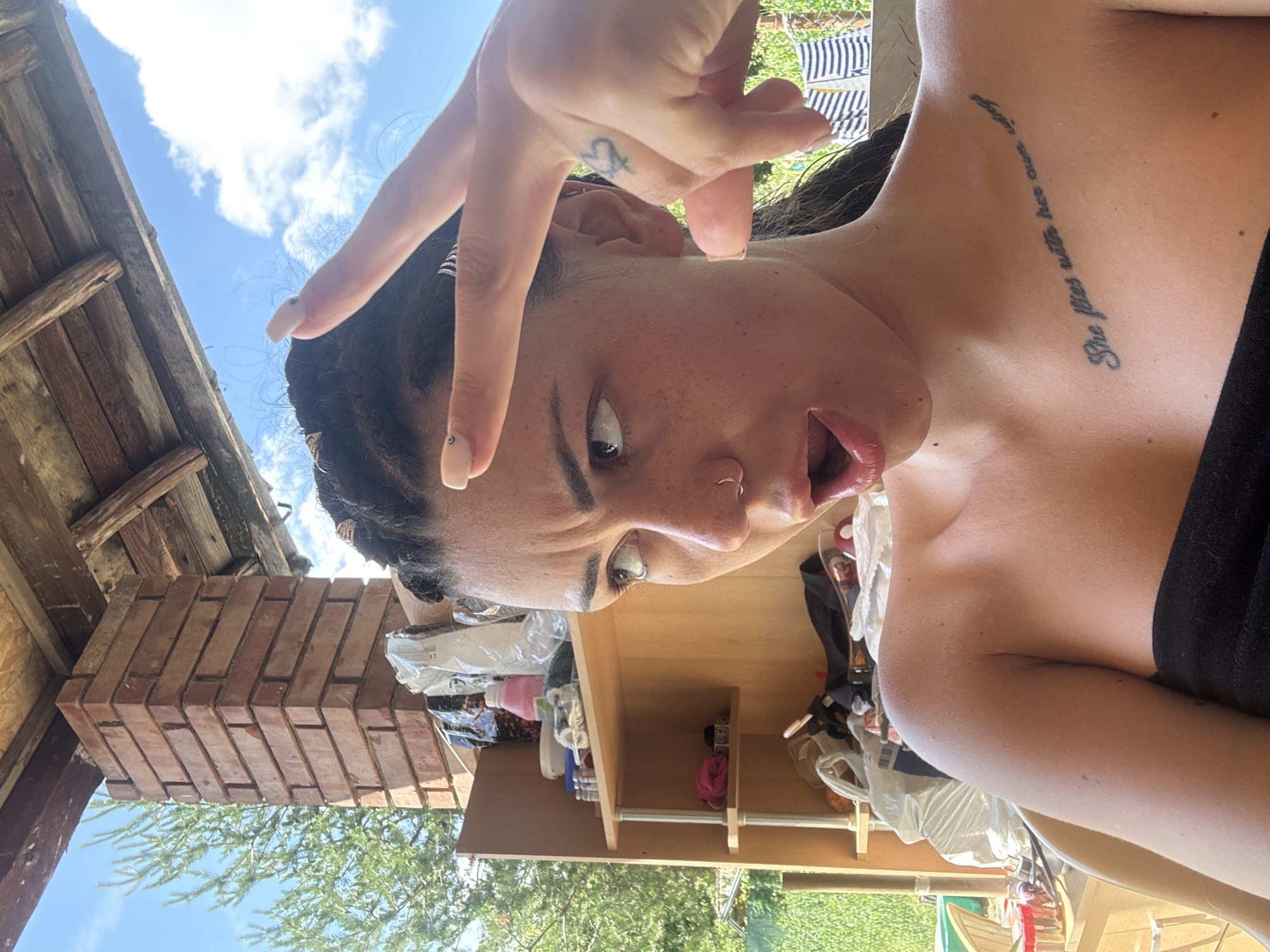 A young woman with braided dark hair, freckles, and a nose ring is posing for a selfie outdoors. She is wearing a black off-the-shoulder top. Her left hand is held up to her face, with two fingers extended in a peace sign. A tattoo of a heart with wings is visible on the back of her right hand. She has a surprised or excited expression with her mouth open.
Behind her, there is a wooden structure with exposed beams and a brick pillar. A shelf unit filled with various items, including bags, bottles, and small containers, is also visible. Green foliage and a blue sky with clouds form the background. The setting appears to be an outdoor area, possibly a patio or porch, with clothes hanging on a line in the distance.
While the image shows an outdoor setting with elements commonly found in a residential area, there is no specific visual information to confirm or deny the location within the city of Belopoptsi, Bulgaria.