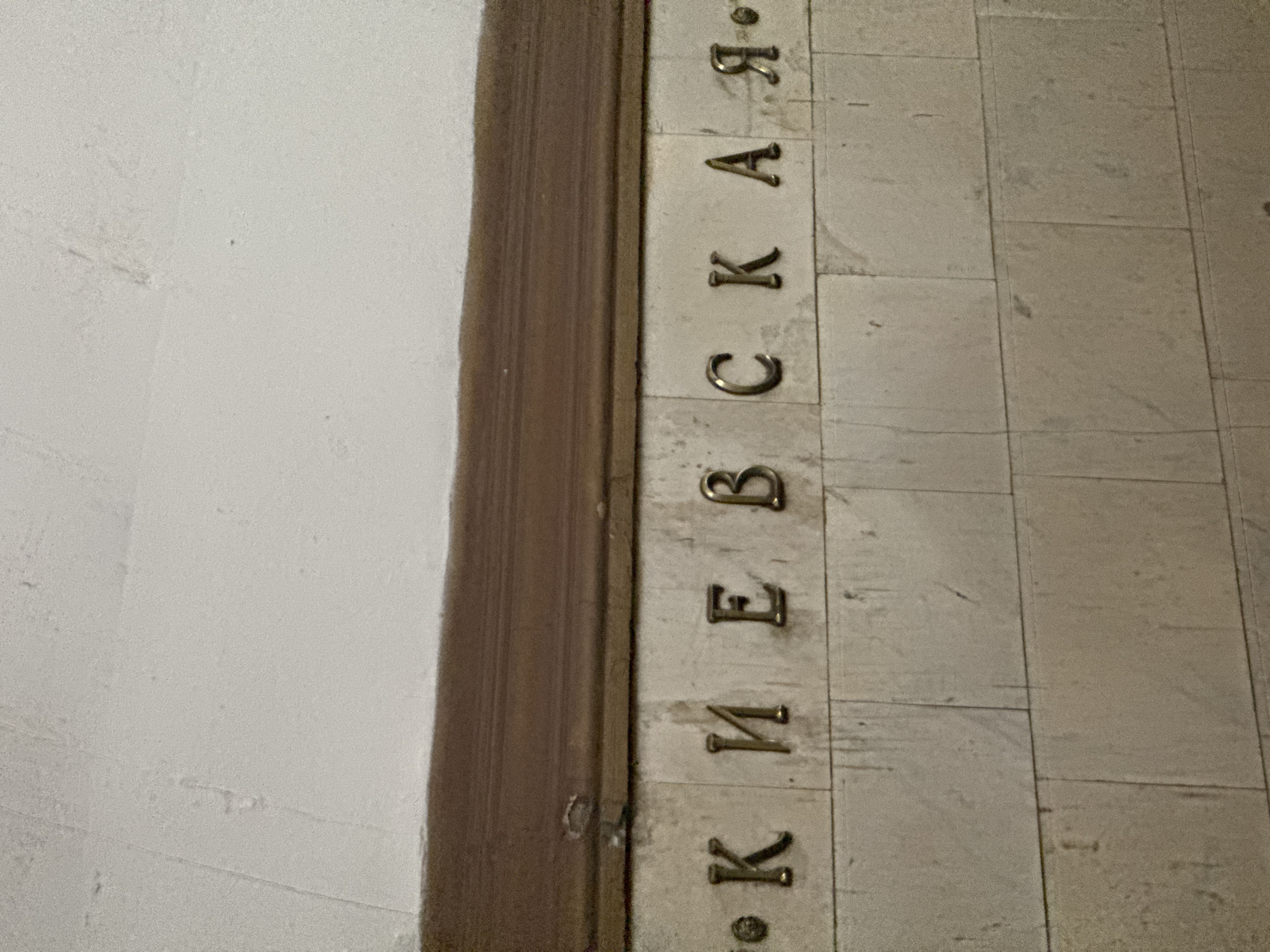 The image shows a sign that reads "Киевская" (Kievskaya) in Russian. The letters are embedded in light-colored stone tiles. The sign is likely located at or near the Kievskaya metro station in Moscow, Russia. A dark wooden frame is visible to the left of the sign. A light gray wall is seen to the left of the wooden frame.