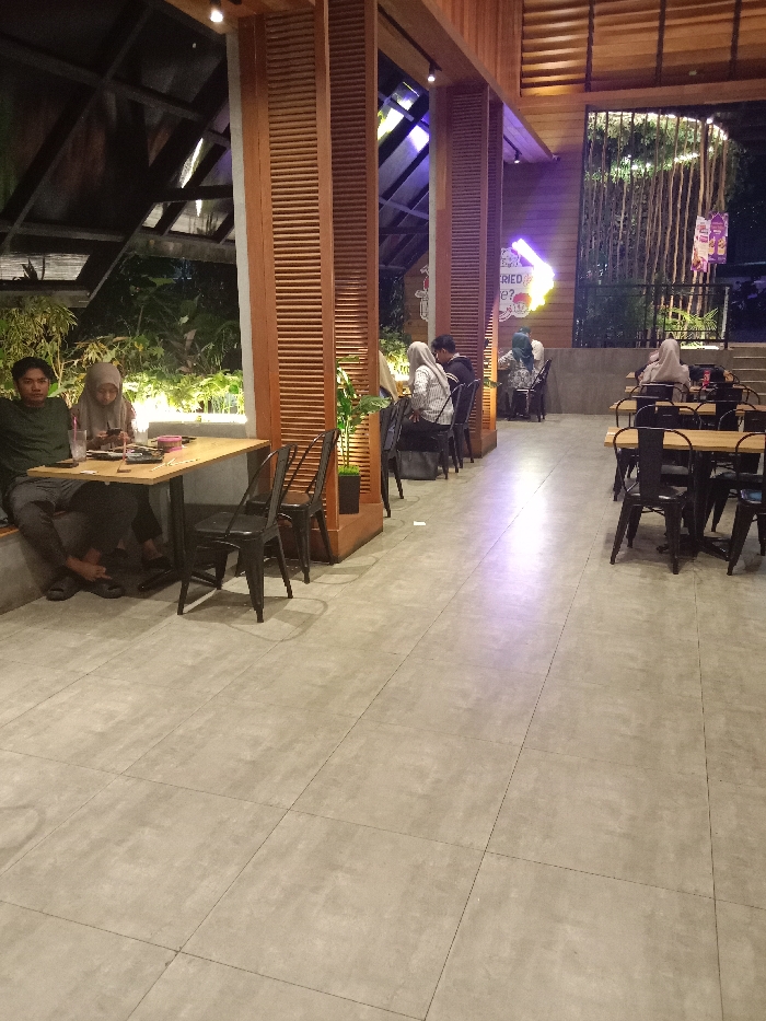 The image shows a restaurant interior with a large open floor space and tiled floor. There are several tables and chairs, some of which are occupied by people. In the foreground, two people are sitting at a table, a man wearing a green shirt and a woman wearing a light pink hijab. They are both looking down at something on the table. In the middle ground, four people are sitting at tables, all facing towards the back of the restaurant. The tables are wooden with black metal chairs. The walls of the restaurant are made of wood and glass, and there are plants in the corners. There are also large windows with views of a courtyard and bamboo plants. The windows are framed with black metal. The image was taken at the coordinates -7.013 latitude and 110.339 longitude, which is located in the Kecamatan Ngaliyan district of Semarang, Indonesia.