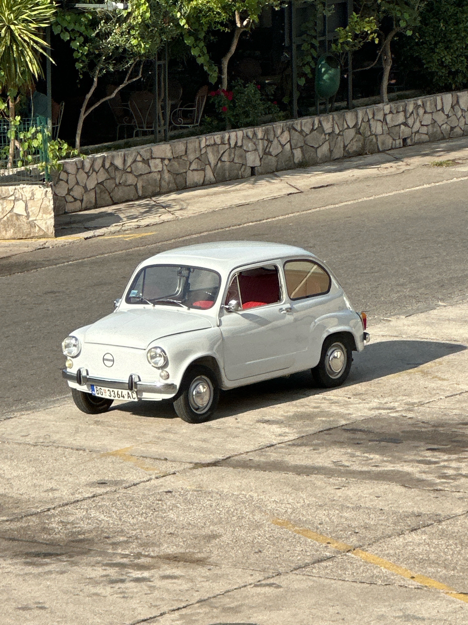 A vintage white Fiat 600 is parked on a concrete surface, possibly a street or parking lot, in Budva, Montenegro. The car is positioned at an angle, facing slightly to the left, with its front end closer to the viewer. It has a bright red interior visible through the windows. The car's license plate reads "BG 3364-AC". Behind the car, a textured stone wall runs horizontally, with lush green foliage and trees growing above it. The scene is illuminated by sunlight, casting a shadow of the car onto the ground. The background shows a hint of a road and more greenery, suggesting an outdoor setting in a town or city.