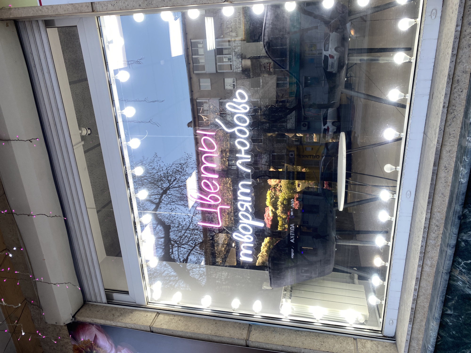 A shop window, likely for a flower shop, is prominently featured, adorned with a bright pink and white neon sign. The sign, written in Cyrillic, reads "Цветы творят любовь," which translates to "Flowers create love." Numerous exposed, bright white light bulbs are arranged vertically along the window's frame, providing additional illumination.
The window's glass acts as a mirror, heavily reflecting a daytime urban environment. Visible reflections include a multi-story building with multiple windows, several bare-branched trees, suggesting autumn or winter, and a parked white car on the street. The sky reflected above is clear and bright.
Additional reflected text from neighboring businesses is discernible. These include "Ателье" (Atelier) and "Срочный ремонт одежды" (Urgent clothing repair), along with "АПТЕКА" (Pharmacy). A partial reflection reading "здравенс" is also visible, possibly part of a larger sign for a health-related service. To the left of the main window frame, a string of small pink decorative lights is partially visible. No people are directly depicted. The scene suggests a clear day in an urban setting, typical of Yekaterinburg, Russia.