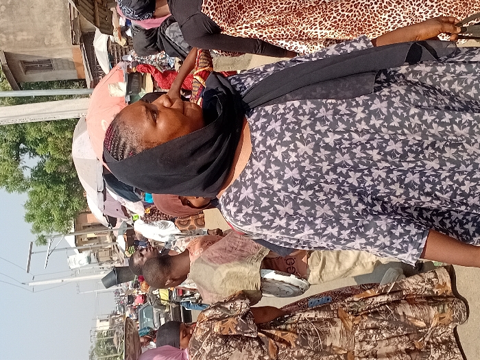 A woman with a dark headscarf and light patterned top is facing left. She is surrounded by other people and various items, likely in a marketplace. Buildings and poles are visible in the background under a bright sky. Another woman wearing a leopard print dress is seen behind the first woman. The scene appears to be outdoors during the daytime.
