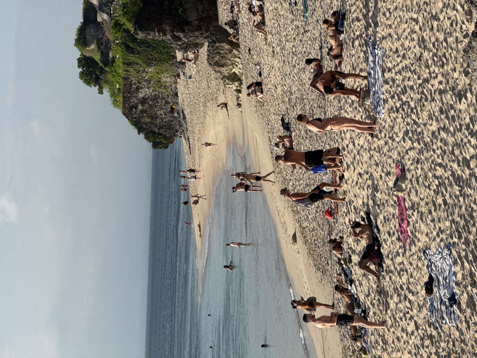The image shows a beach scene in Kuta Selatan, Indonesia. The beach is sandy and crowded with people sunbathing, swimming, and walking along the shore. A cliff with green vegetation rises on the left side of the beach. The water is clear and blue, and the sky is hazy.