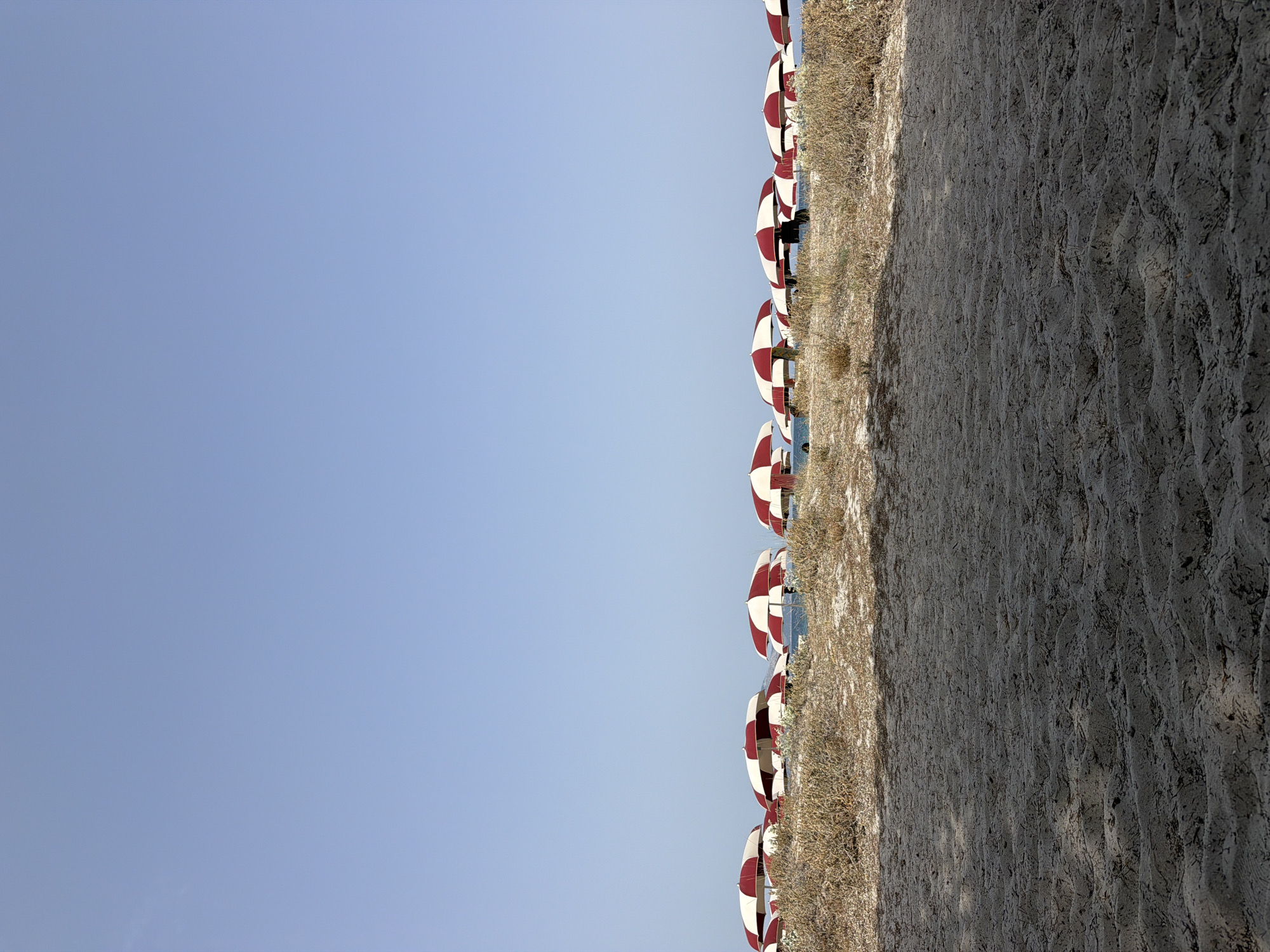 The image depicts a beach scene in Olbia, Italy, on a clear day. A row of red and white striped beach umbrellas stretches across the middle ground, casting shadows on the dry, sandy ground and sparse vegetation. The sand in the foreground is textured with ripples and debris. The sky above is a uniform, pale blue. No people are clearly visible. The umbrellas appear to be set up for rental, suggesting a public beach.
