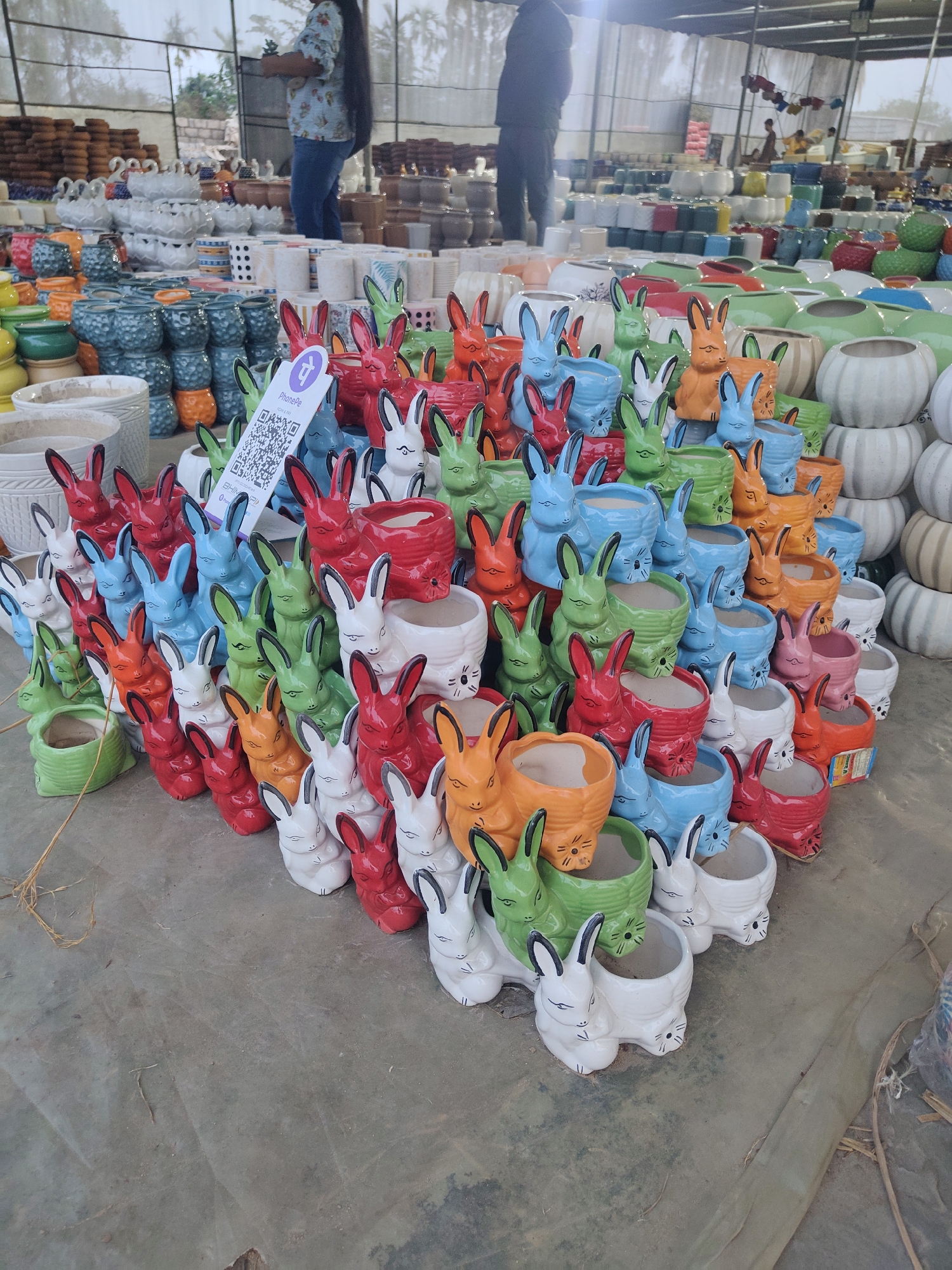 The image depicts a large collection of rabbit-shaped ceramic planters in various colors, stacked in a pyramid formation. The planters are arranged on a concrete surface, likely in an outdoor setting. In the background, there is a wide array of other ceramic pots and containers. Some people are visible in the background, appearing to be shopping or working in the area. There is also a QR code displayed on one of the planters. The presence of the QR code, the diversity of ceramic planters, and the background activities indicate a market or retail setting, possibly in the city of ಚಂದ್ರಪ್ಪ ವೃತ್ತ, India.