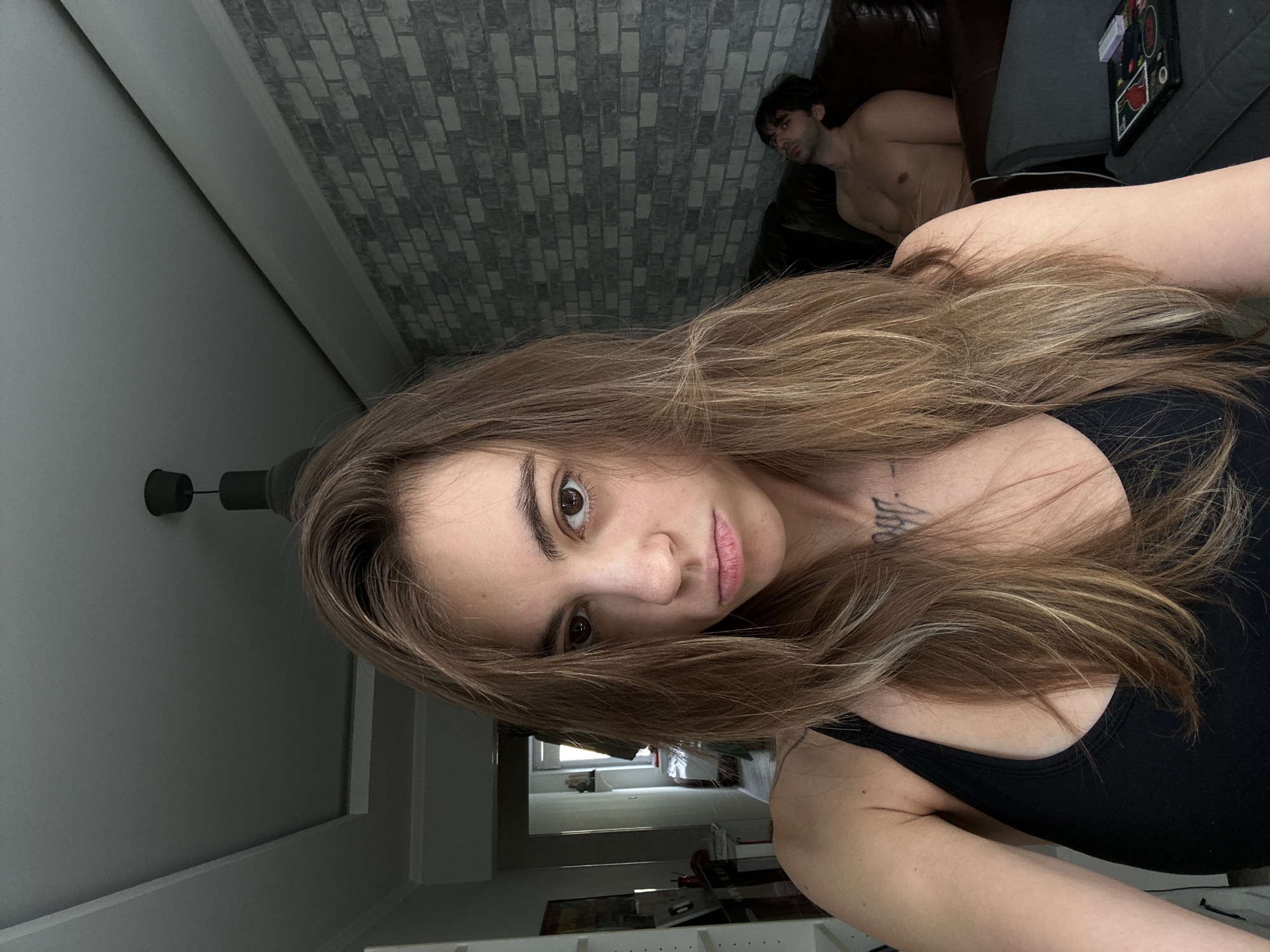 A woman is taking a selfie in a room, with a man lying on a couch in the background. The woman is in the foreground, looking at the camera with a pout. She has long, light brown hair and is wearing a black tank top. There is a tattoo on her chest. The man in the background is shirtless and appears to be sleeping or resting. He is lying on a dark brown leather couch. The room has a modern design with gray brick wallpaper and a textured ceiling. A dark pendant light hangs from the ceiling. To the left of the woman, a hallway is visible, with shelves and what appears to be a door. On the right side of the room, another piece of furniture, possibly a chair or ottoman, holds a tablet device and some small items. The lighting suggests it might be daytime, but the room is relatively dim. The image appears to be taken indoors, likely in an apartment. Without further information, it is not possible to determine the specific location within Moscow, Russia.