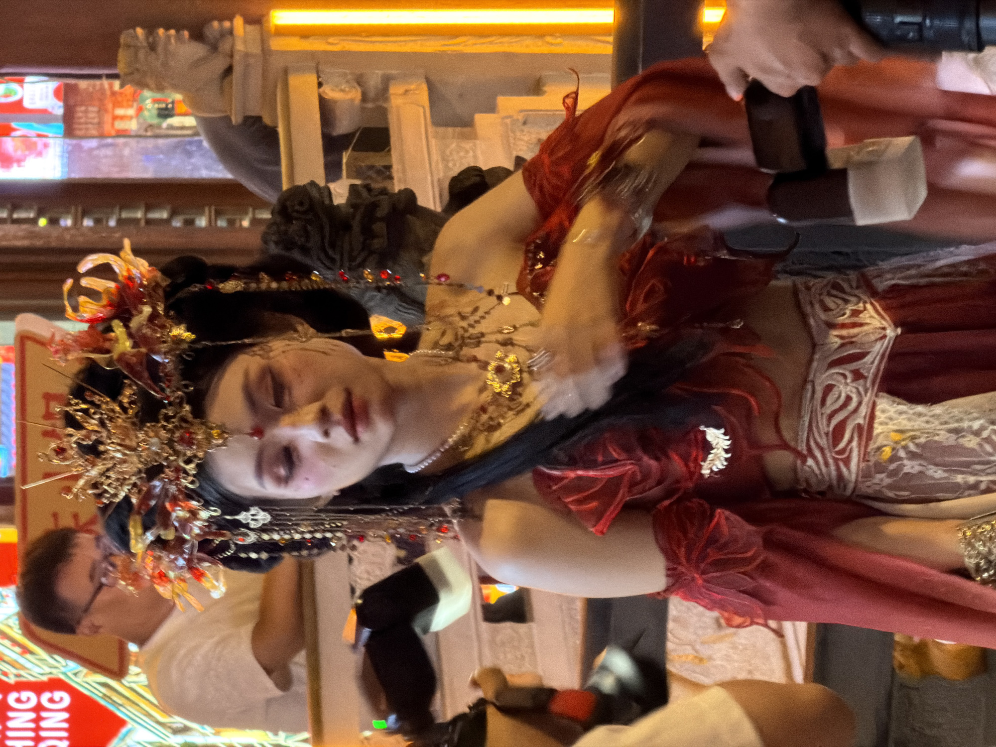 A young Asian woman in elaborate traditional Chinese attire, featuring a gold headdress with red and gold accents and a red off-the-shoulder dress with sheer red sleeves, stands center frame. She has dramatic makeup with pink blush and eye shadow. Behind her, a man wearing glasses and a white t-shirt is visible. The background suggests an indoor setting with decorative elements like stone lion statues and illuminated signage with Chinese characters. A person's arm holding a mobile phone is visible in the lower right corner. The overall scene has warm, artificial lighting.