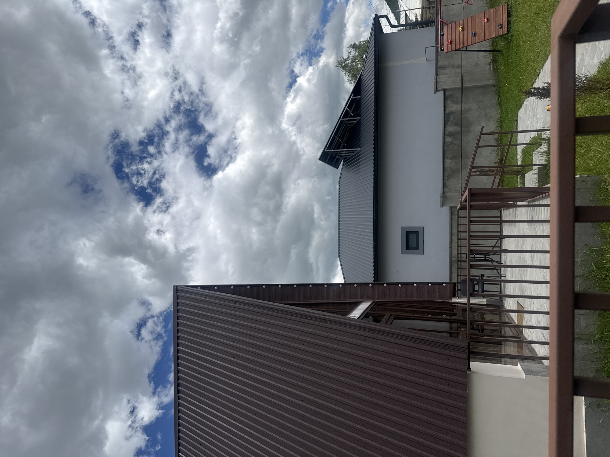 A low-angle shot captures the exteriors of two adjacent buildings under a partly cloudy sky in Stepanavan, Armenia. The building in the foreground features a brown corrugated metal roof, which dominates the lower left portion of the frame. A glimpse of its light-colored exterior wall is visible.
The adjacent building, partially visible, has a gray corrugated metal roof and a light gray exterior wall. A small, square window is set into the wall. To the right of this building, there is a raised outdoor area with a brown railing and steps leading down. A small section of grass is also visible. Further back, a structure resembling a climbing wall is present.
The sky is filled with a mix of gray and white clouds, with patches of blue sky visible in between. The overall impression is of a quiet, possibly residential or commercial area, with modern building designs.
