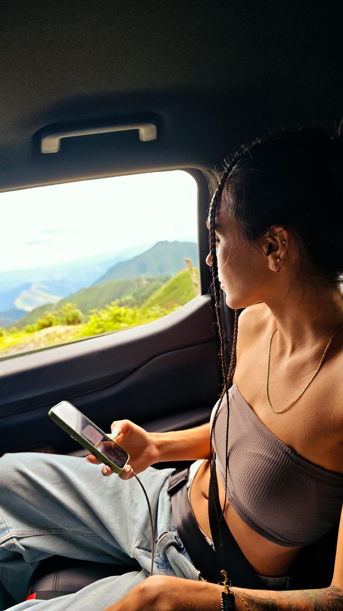 A young adult female is seated in the rear passenger side of a vehicle, looking out a window at a mountainous landscape in Xín Vàng, Vietnam. Her left profile is visible as she gazes towards the verdant mountains under a bright, clear sky. She has dark hair styled in micro-braids, pulled back, with loose strands framing her face. She wears multiple ear piercings, a delicate gold chain necklace, and a dark grey ribbed strapless crop top. Her lower body is clad in light blue denim jeans, and a black seatbelt is fastened across her lap. In her left hand, she holds a smartphone with a prominent green case, connected by a dark charging cable that extends downwards. The phone screen is dark and reflective. The vehicle's interior features a dark, textured headliner and a light-colored overhead grab handle. A dark door panel is visible below the window. Outside the window, multiple layers of green mountains, some appearing cultivated, recede into a hazy blue distance under daylight conditions.