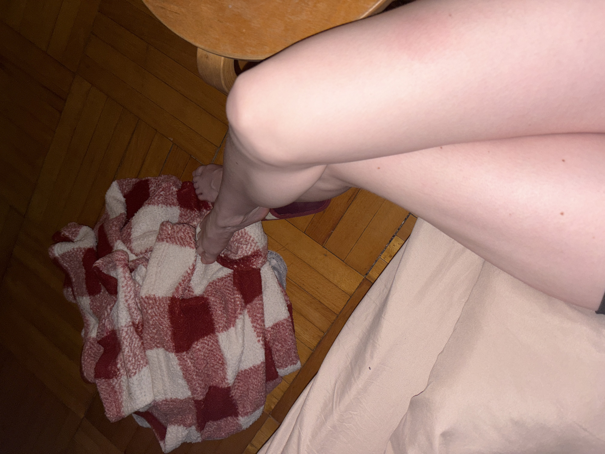 The image shows a person's legs crossed under a wooden table. A red and white checkered blanket is bunched up next to the legs. A portion of a light pink bedsheet is visible on the right side. The floor is made of wooden planks.