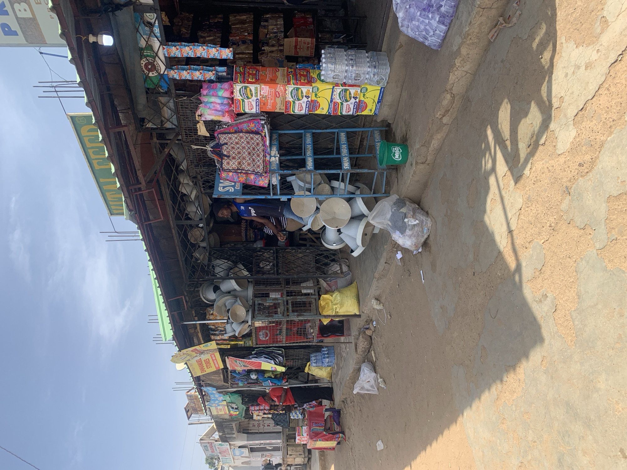 Here is a forensic description of the image:
**Setting:** The image depicts an outdoor street scene in Rafin Par, Nigeria. The surface is rough, sandy, and uneven. A shadow is cast on the ground.
**Scene Description:** The primary focus is a roadside shop or stall crammed with various goods. The stall has a metal frame and is covered with a green corrugated roof. Numerous signs are visible, including those for "Emmanuel Stores", "MEEK PHARMACE", "SV", "Indutras", "MILO", and "PPC". Merchandise is stacked on shelves and hanging from the structure. These items include:
* Packaged food (possibly noodles, pasta, and milk)
* Bottled water
* Pots and pans (aluminum or stainless steel)
* Bags of unknown contents
* Clothes or fabric
**People:** A man is visible behind the stall, wearing a blue soccer jersey. It is unclear exactly what he is doing.
**Objects:** Other objects visible include a green bucket marked "MILO," miscellaneous bags and containers, and an electrical lightbulb hanging from the stall's roof.
**In summary:** The image captures a typical roadside commercial scene in Rafin Par, Nigeria, characterized by a cluttered storefront offering a variety of consumer goods. A man stands behind the stall.