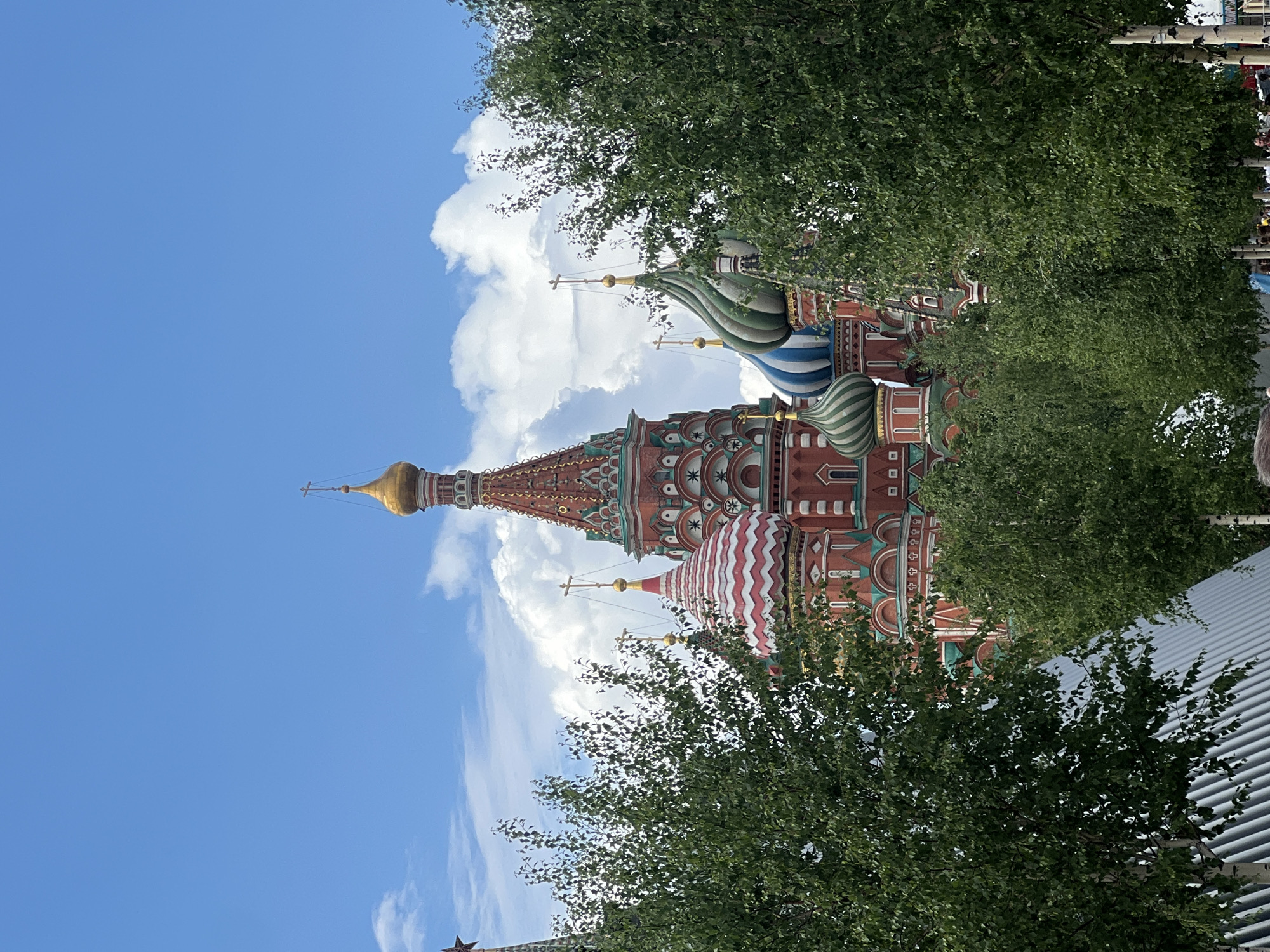 The image depicts the iconic Saint Basil's Cathedral in Moscow, Russia. The cathedral's distinctive colorful domes and spires rise against a backdrop of blue sky and white clouds. Trees with green foliage partially obscure the view of the building.