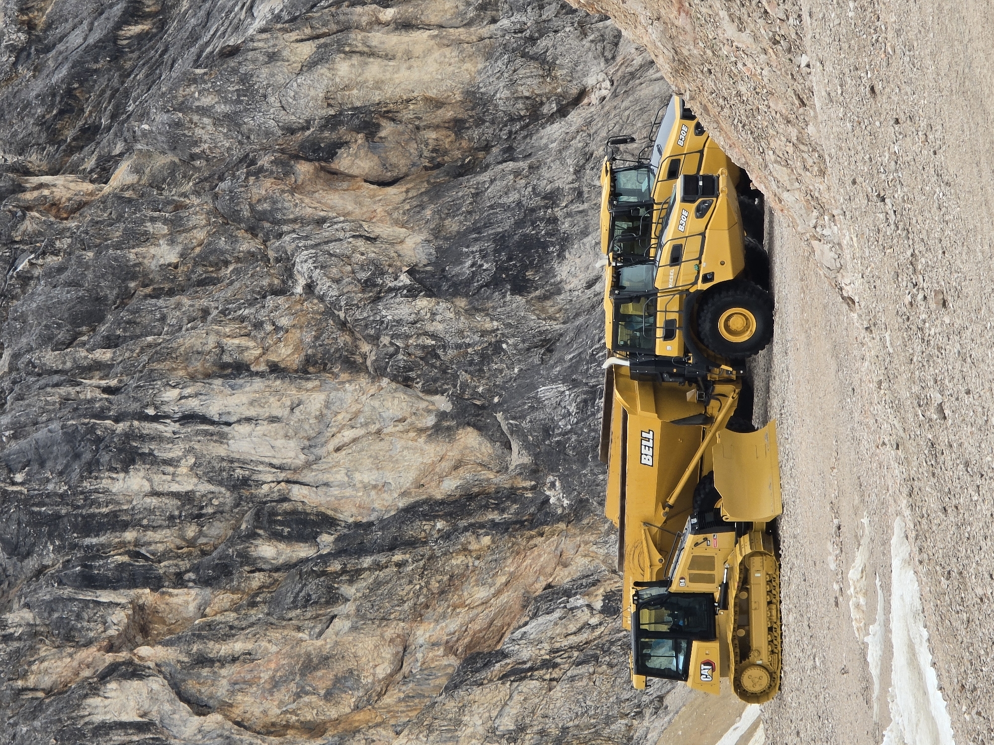 A line of yellow construction vehicles is parked against a steep, rocky mountainside. The vehicles include a Caterpillar bulldozer and at least two Bell articulated dump trucks. The bulldozer has "CAT" visible on its side. The dump trucks have "BELL" visible on their bodies. One of the dump trucks has "B30E" visible on its side. The terrain is rugged and appears to be a mine or construction site. The weather is clear and the time of day is likely daytime, given the lighting. The scene suggests heavy-duty construction or mining operations are taking place in a mountainous region, potentially in Garmisch-Partenkirchen, Germany, as indicated by the location context.