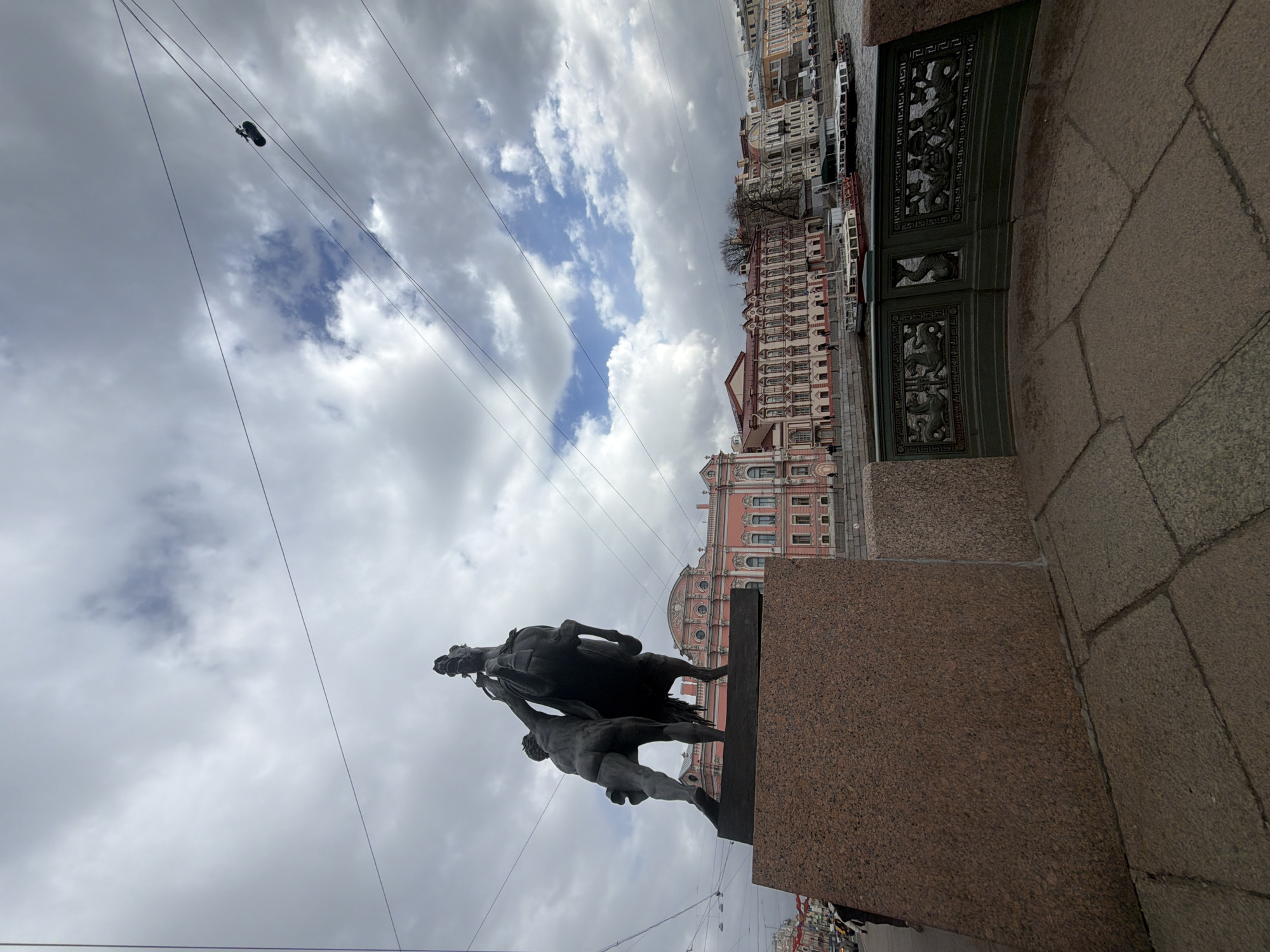The image depicts a statue in Saint Petersburg, Russia, positioned on a granite pedestal. The statue portrays a human figure struggling to control a horse. In the background, ornate buildings line a canal. The sky is filled with clouds, with a small patch of blue visible. Overhead, wires with suspended objects, potentially shoes, stretch across the sky. A decorative metal fence is partially visible to the right of the frame.