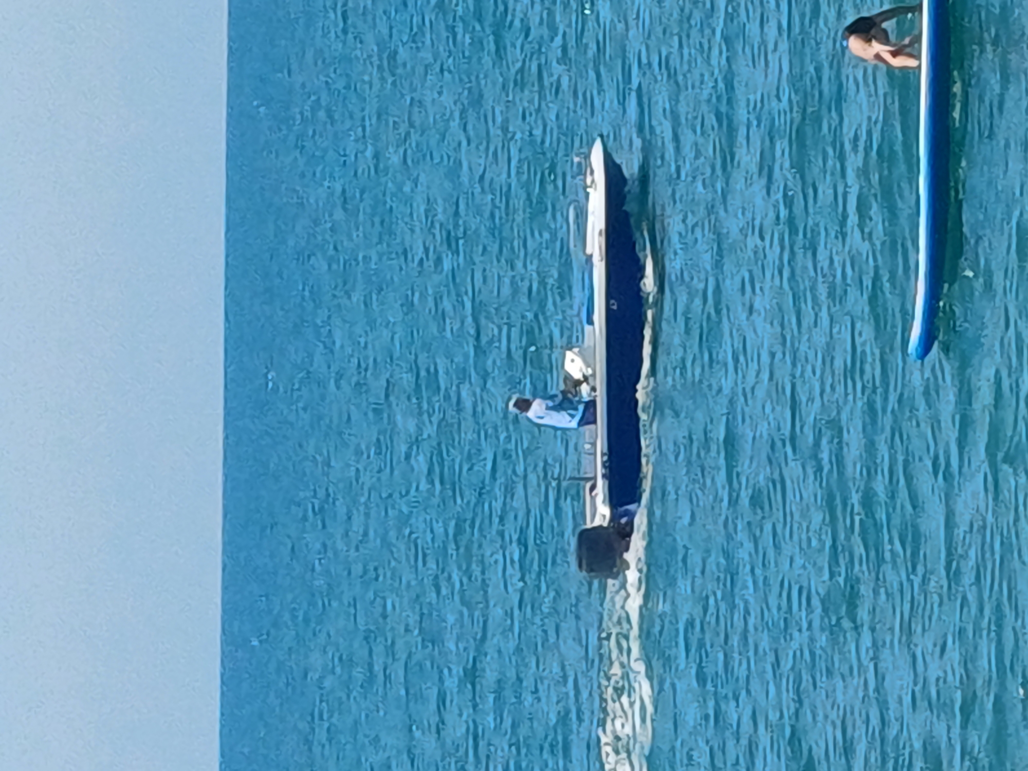 A person is operating a small motorboat on a body of water. The boat is white with blue accents and has an outboard motor. The person is wearing a light-colored shirt and a hat. Another person is on a blue paddleboard in the foreground, attempting to stand. The background shows a vast expanse of blue water and a light blue sky. The scene is likely a coastal area, potentially in Sochi, Russia, given the clear, calm waters and recreational activities depicted.