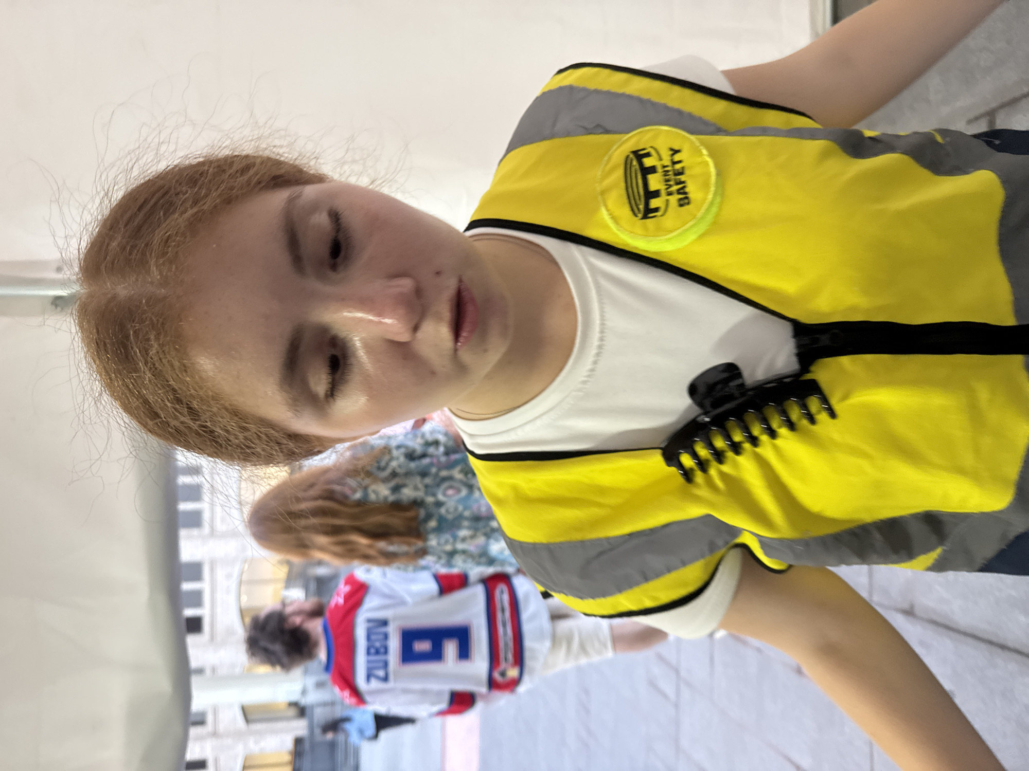 A close-up, medium shot shows a young woman with light brown hair wearing a bright yellow safety vest over a white t-shirt. The vest has grey reflective stripes. A black comb is clipped to the vest on the left side. A circular yellow patch with the words "EVENT SAFETY" is visible on the right side of the vest. Her eyes are partially closed, and her mouth is slightly open. In the background, slightly out of focus, a man wearing a white hockey jersey with "ZUBOV" and the number "9" on the back is walking away from the camera. Another person with long red hair is also visible behind him. The background appears to be an outdoor urban setting with architectural elements suggesting a large building. The lighting indicates it is daytime. No specific location in Moscow can be identified from the image.