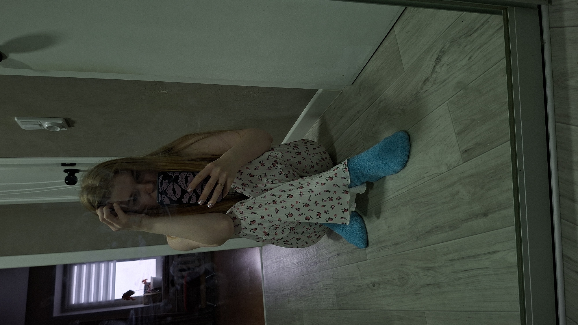 The image, rotated 90 degrees clockwise from its natural orientation, depicts a person captured in a mirror reflection within an indoor setting. A person with long, light-brown hair is shown seated on a light wood-patterned floor, holding a smartphone in their left hand, while their right hand rests near their face, partially obscuring it.
The person is wearing light-colored pajamas or loungewear with a repetitive small floral pattern in shades of red or pink, paired with bright blue, fluffy socks. Their fingernails are visibly manicured. The smartphone case is dark, featuring a pattern of lighter, abstract shapes, possibly lips or footprints.
The background, visible through the mirror's reflection, includes a light-colored wall or ceiling with a white light fixture. Below this, a dark door frame and handle are partially visible. A window with horizontal blinds is also reflected, indicating a source of ambient light. The mirror itself has a thin, light-colored frame along its right edge. The overall lighting is subdued, creating a calm atmosphere. The pose, with a hand near the face, suggests a casual or perhaps contemplative moment. The location is Chaikovsky, Russia.