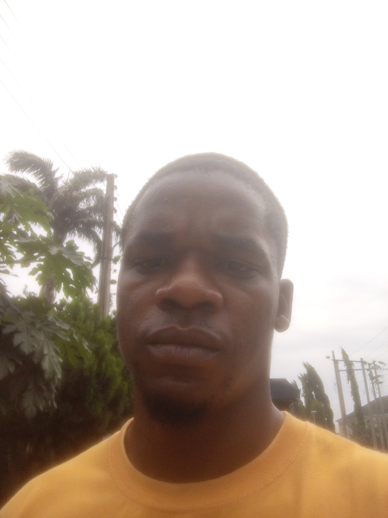 A selfie taken in Gwarinpa, Nigeria, shows a dark-skinned man with short hair. He is wearing a yellow t-shirt. The background is overcast, with some trees visible, including what appears to be a palm tree, and power lines. The lighting suggests it is daytime.