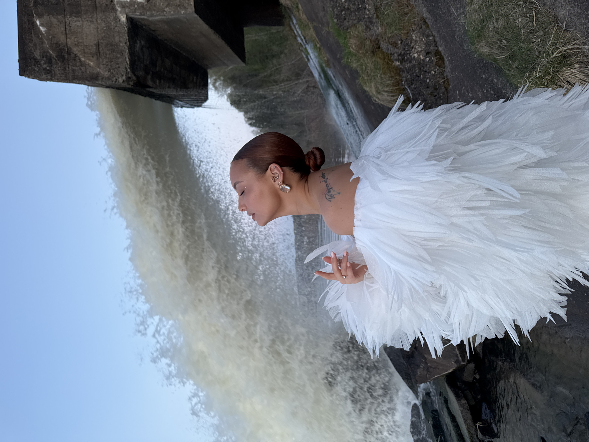 A woman is standing in front of a waterfall on what looks like the side of a dam in Noviy, Russia. The woman has brown hair styled in a bun, light skin, and is wearing earrings. She has a tattoo on her neck. She is wearing a white garment made of feathery or fringed material that covers her shoulders and possibly extends into a dress. She is looking down at the water, with her right hand slightly raised. The waterfall is in the background, with the dam structure visible above. The sky is light blue. There is some vegetation growing on the side of the dam. The photograph is oriented vertically.