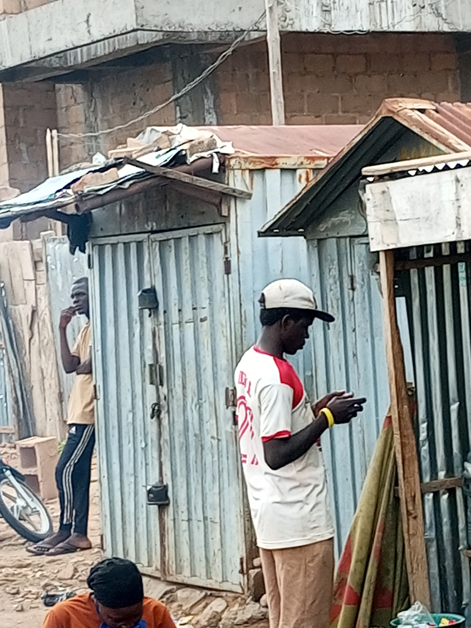 In Tempure, Nigeria, two young men are visible in a street scene. The man on the left, facing away from the camera, stands near a corrugated metal structure, wearing a tan t-shirt and black pants with white stripes. He has his hand near his mouth. To the right, another young man, facing the camera from the side, wears a white t-shirt with red sleeves, tan pants, and a light-colored baseball cap. He is looking down at his mobile phone, which he holds in his hands, and is wearing a yellow wristband. The environment consists of makeshift structures made of corrugated metal and what appears to be a partially constructed building in the background. A bicycle wheel is visible on the left side of the frame. The ground is covered in dirt and debris.