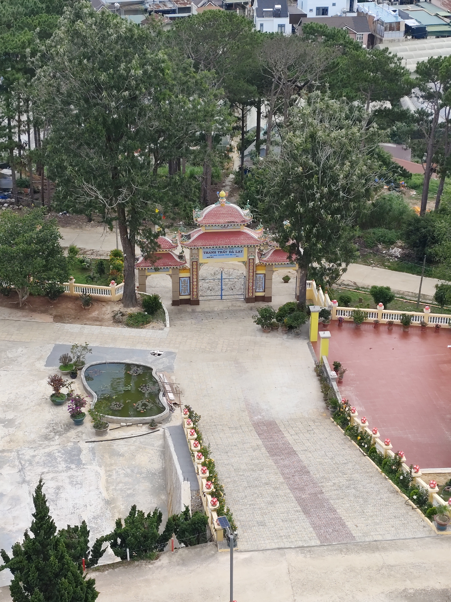 The image captures an aerial view of a temple complex in Lộc Quý, Vietnam. The central focus is a traditional temple gate with a red-tiled roof, adorned with ornate yellow and blue decorations. Beyond the gate, a courtyard is paved with patterned tiles, and a large, red-tiled area is visible to the right, likely an outdoor space for gatherings or ceremonies.
To the left of the gate, a low, ornate yellow fence encloses a garden area with trees and bushes. In the foreground, a heart-shaped pond filled with green water and lotus plants adds a serene element to the scene. Various potted plants are strategically placed throughout the complex, enhancing the greenery. A tall pine tree stands prominently near the gate, casting a shadow over the paved courtyard.
The surrounding environment is characterized by lush vegetation, including numerous trees and dense foliage, suggesting a semi-rural or well-landscaped urban setting. Residential buildings and greenhouses are visible in the background, indicating the presence of a community nearby. The overall atmosphere appears peaceful and well-maintained, typical of a religious or cultural site. There are no people visible in the image.